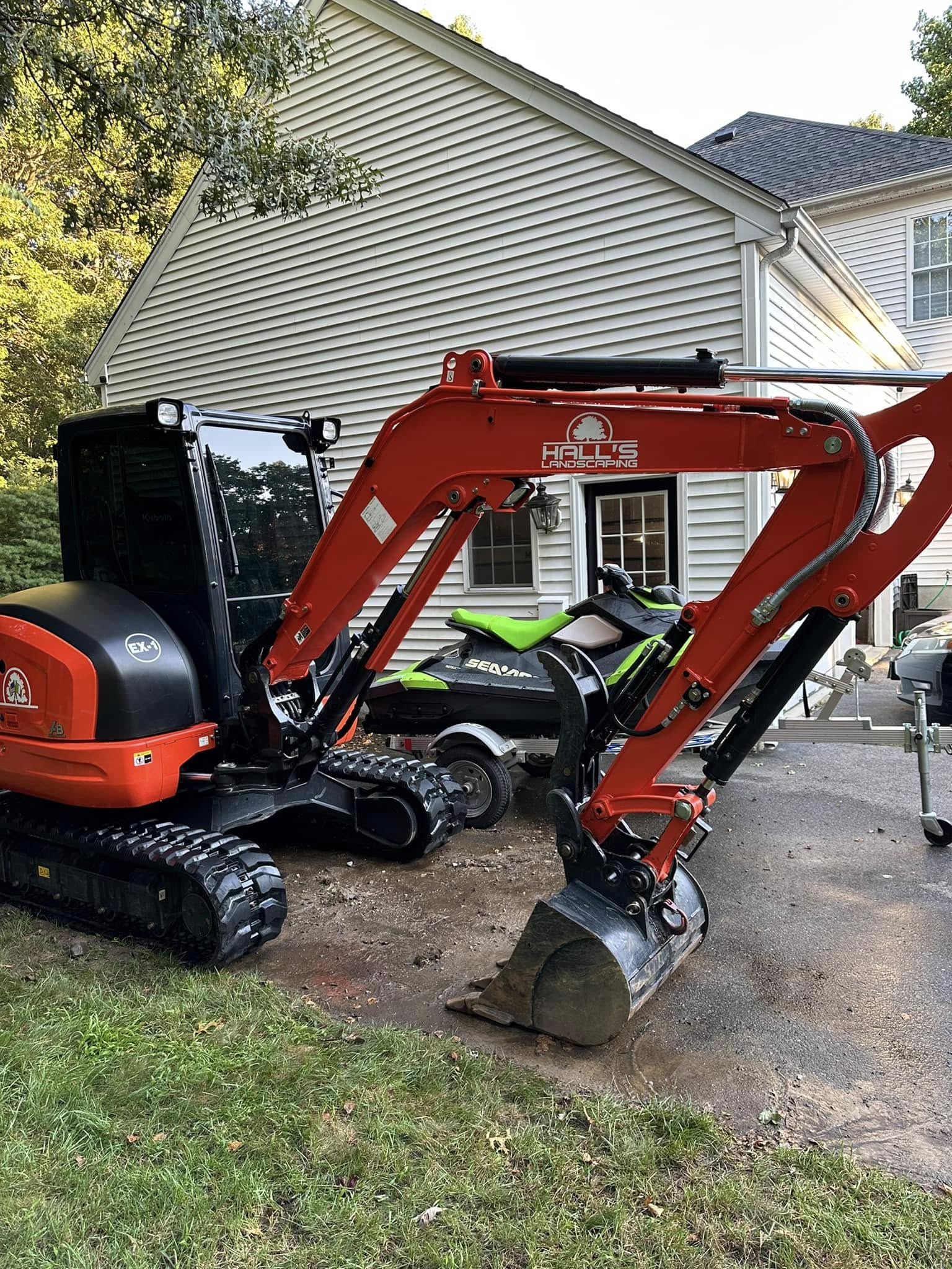 Red mini excavator on a gravel driveway beside a house with a black dumpster and trees nearby