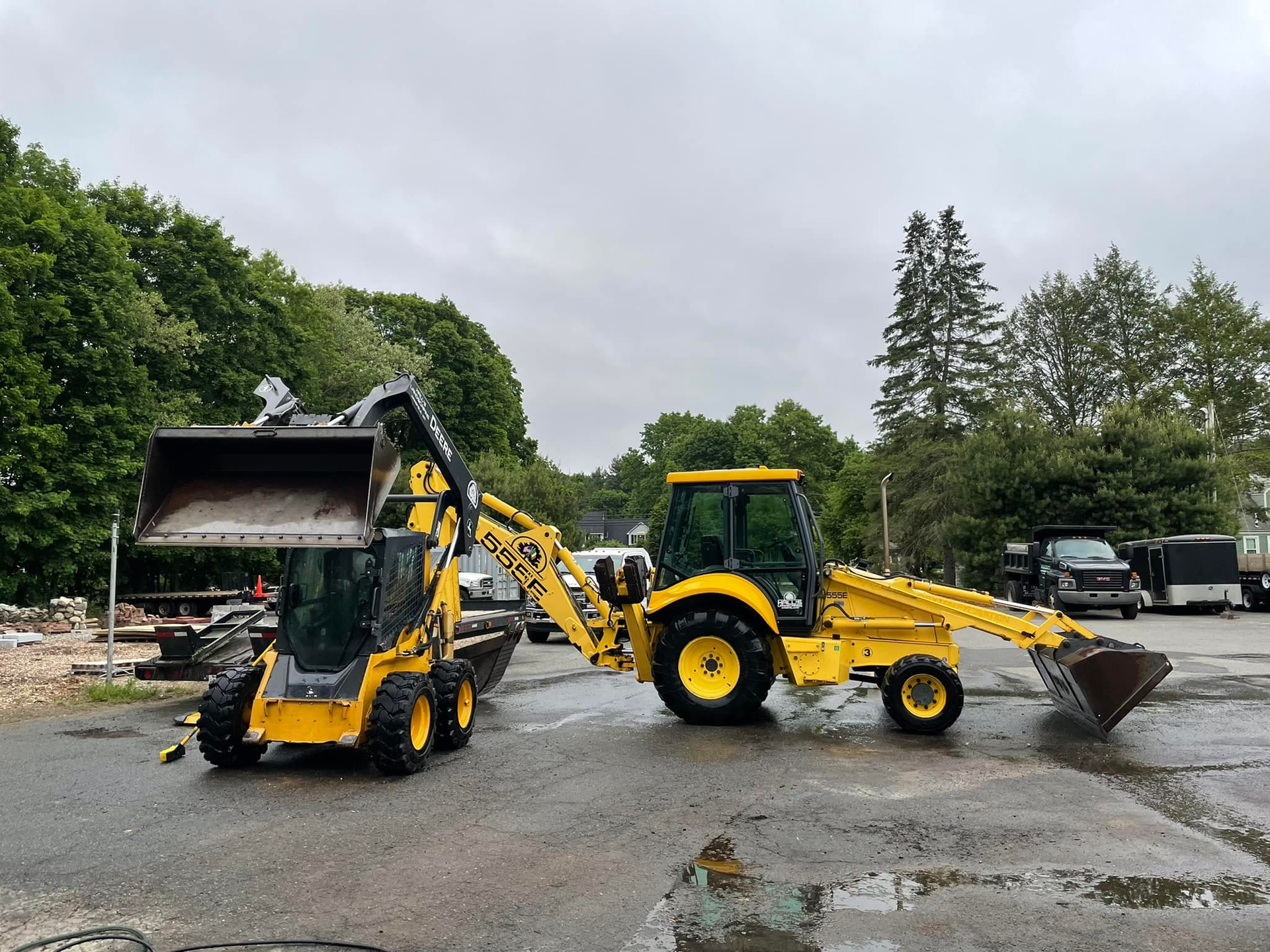 Yellow backhoe loader beside an overturned construction vehicle in a muddy lot under cloudy skies