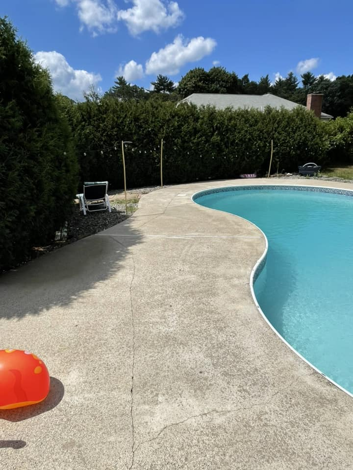 Curved backyard swimming pool with concrete deck, lounge chair, hedge, and blue sky with clouds