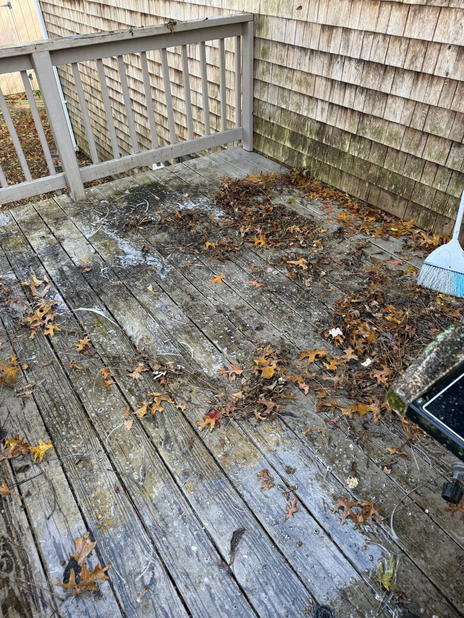 Weathered wooden deck with wet leaves, dirt, and debris beside a shingled wall and railing.