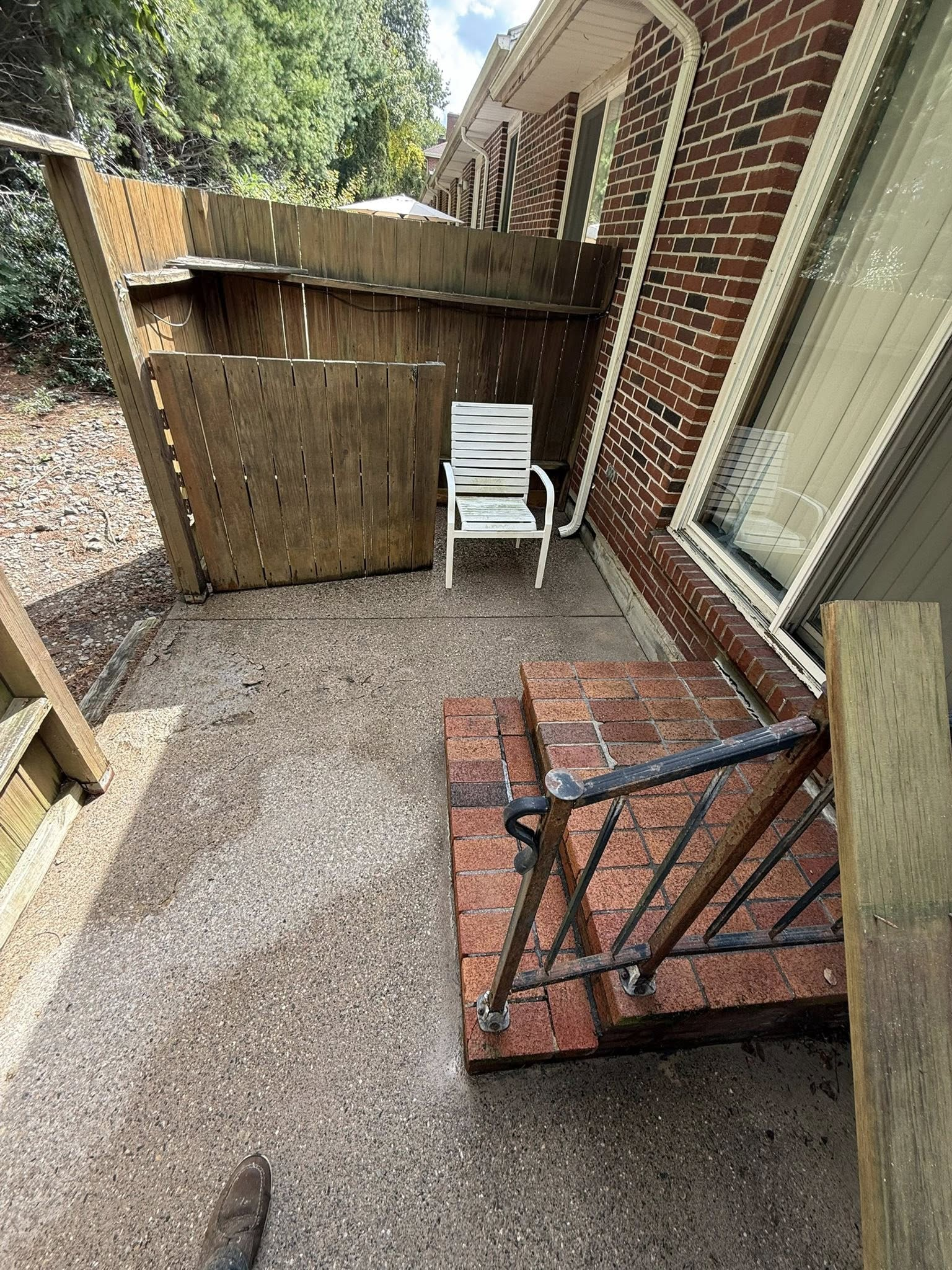 Narrow brick patio with gravel, a white chair by a wooden fence, and a brick step with black railing.