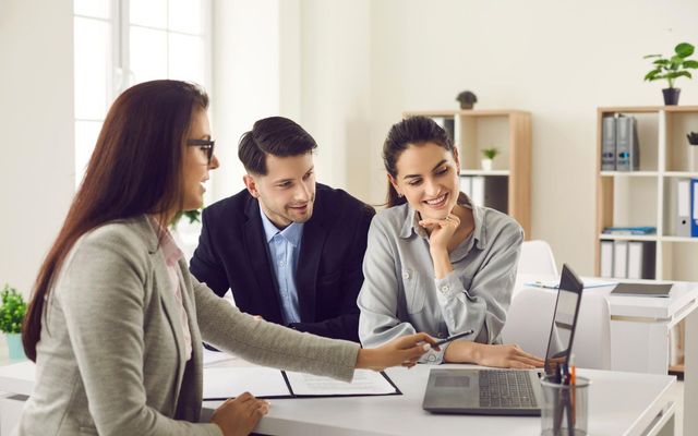 Three coworkers in an office discussing papers around a desk with a laptop and whiteboard in the background