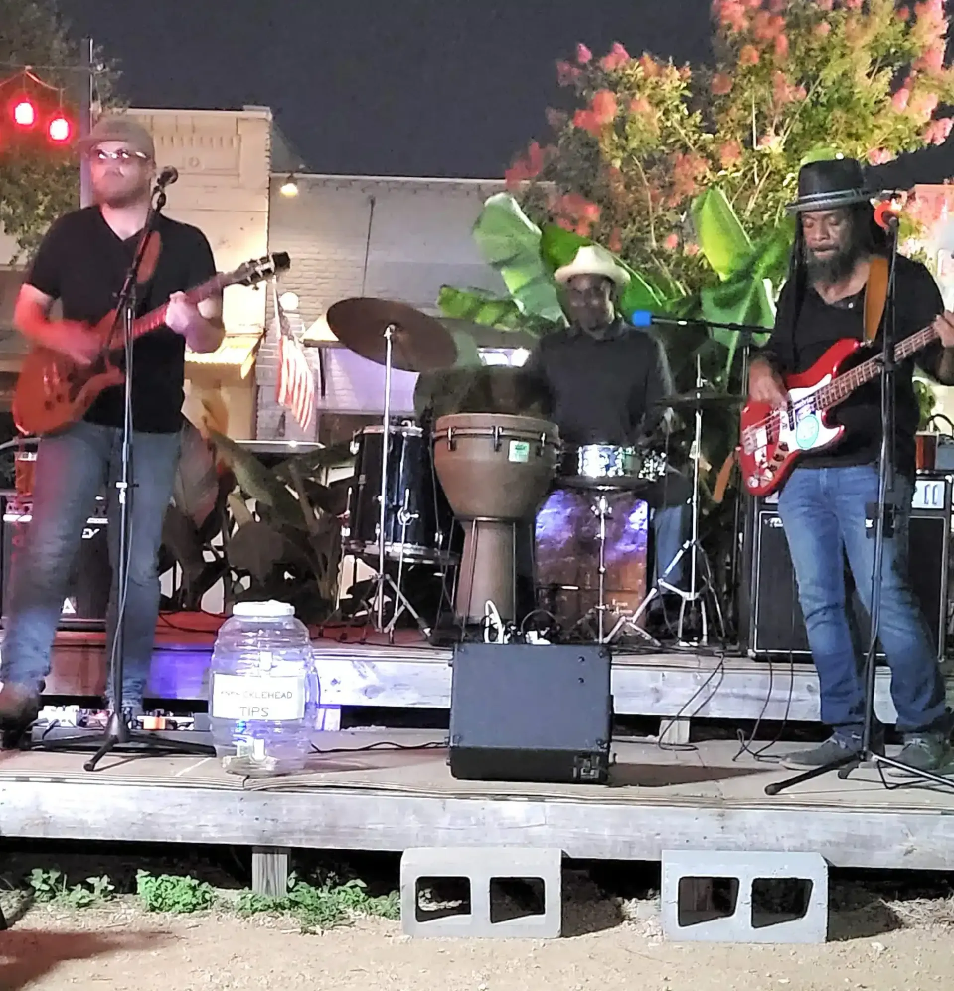 Outdoor band performing on a small stage at night, with guitars, drums, and colorful lights.