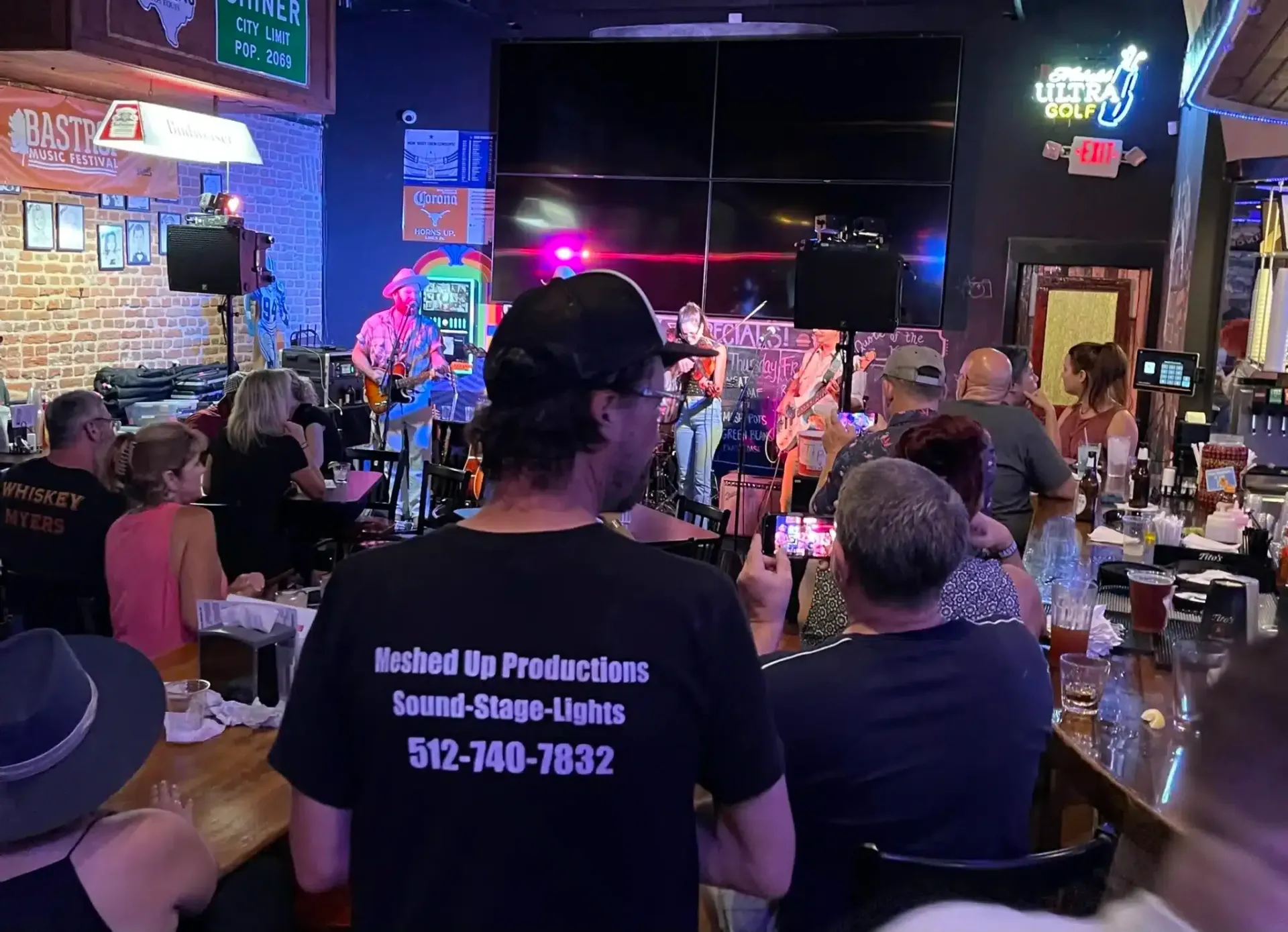 Crowded bar with live music, neon signs, and patrons watching a performance at tables and stools
