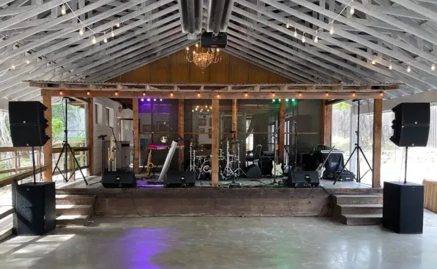 Empty indoor stage with instruments and speakers under a wooden roof, lit by purple lights