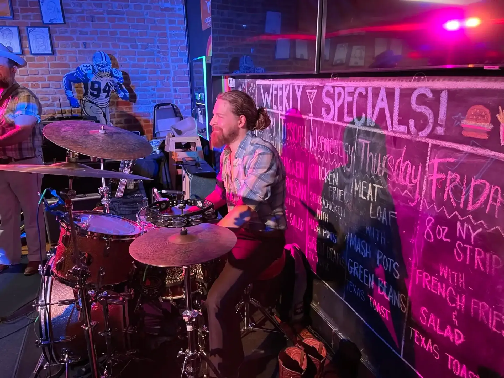 Drummer performing onstage under pink and blue lights beside a chalkboard specials sign