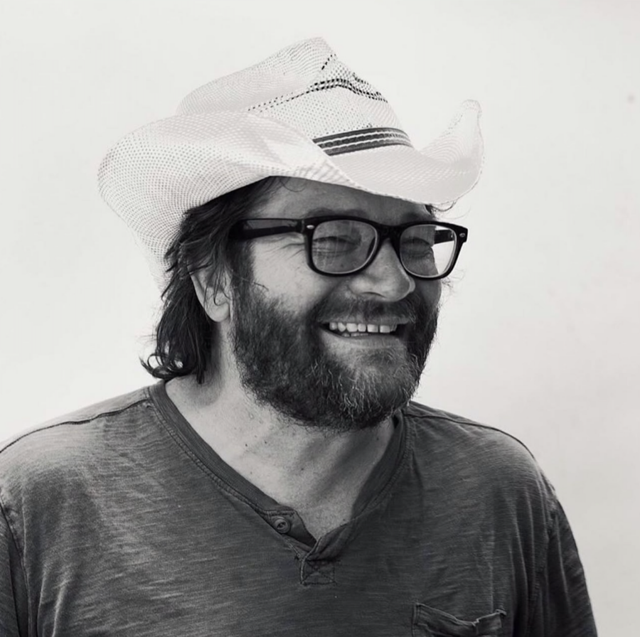 Smiling person wearing glasses and a white cowboy hat on a plain background