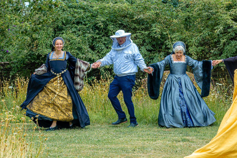 A man in a beehive is holding hands with two women in Tudorcostumes.