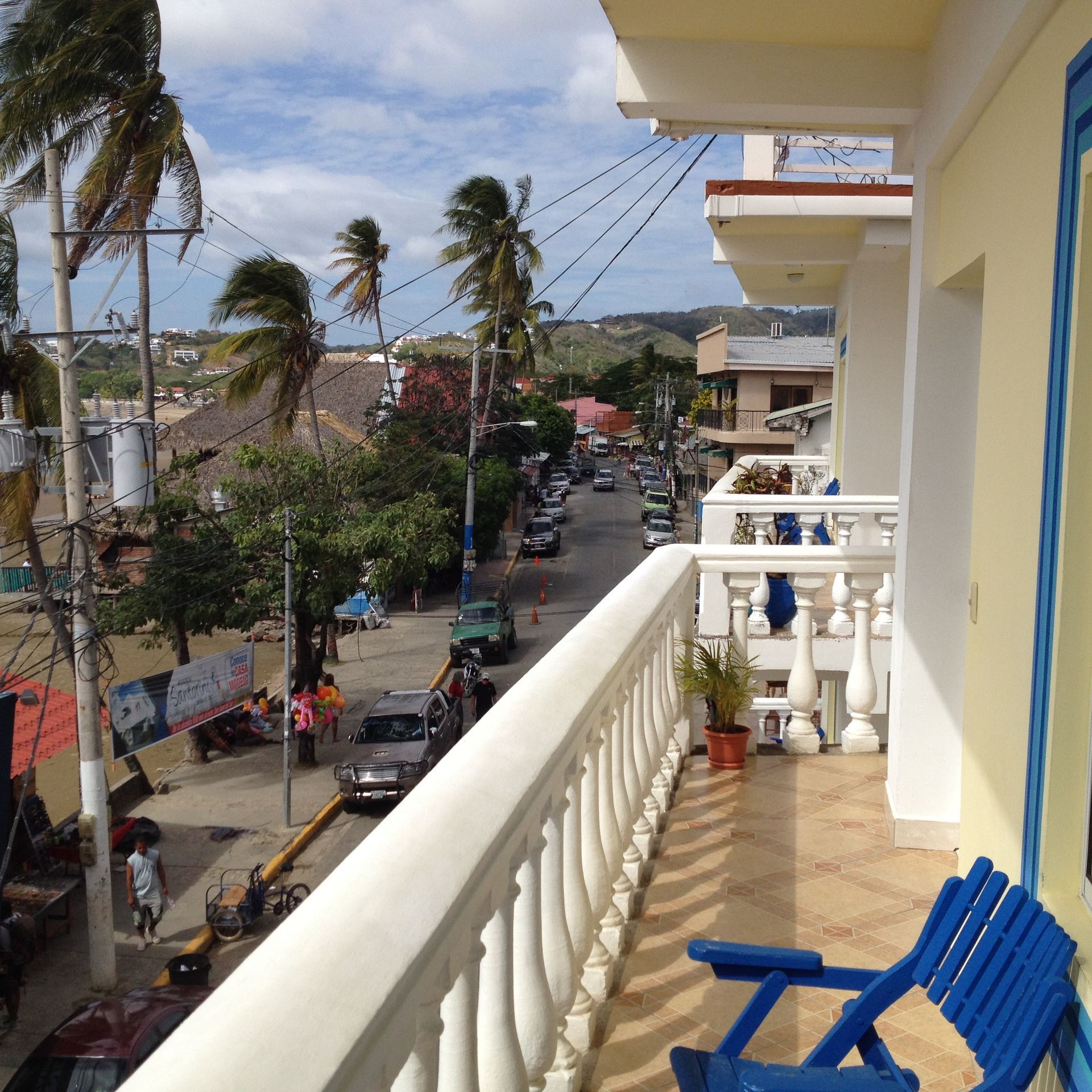 View from a balcony overlooking a street with cars, palm trees, and buildings; blue chair on the balcony.