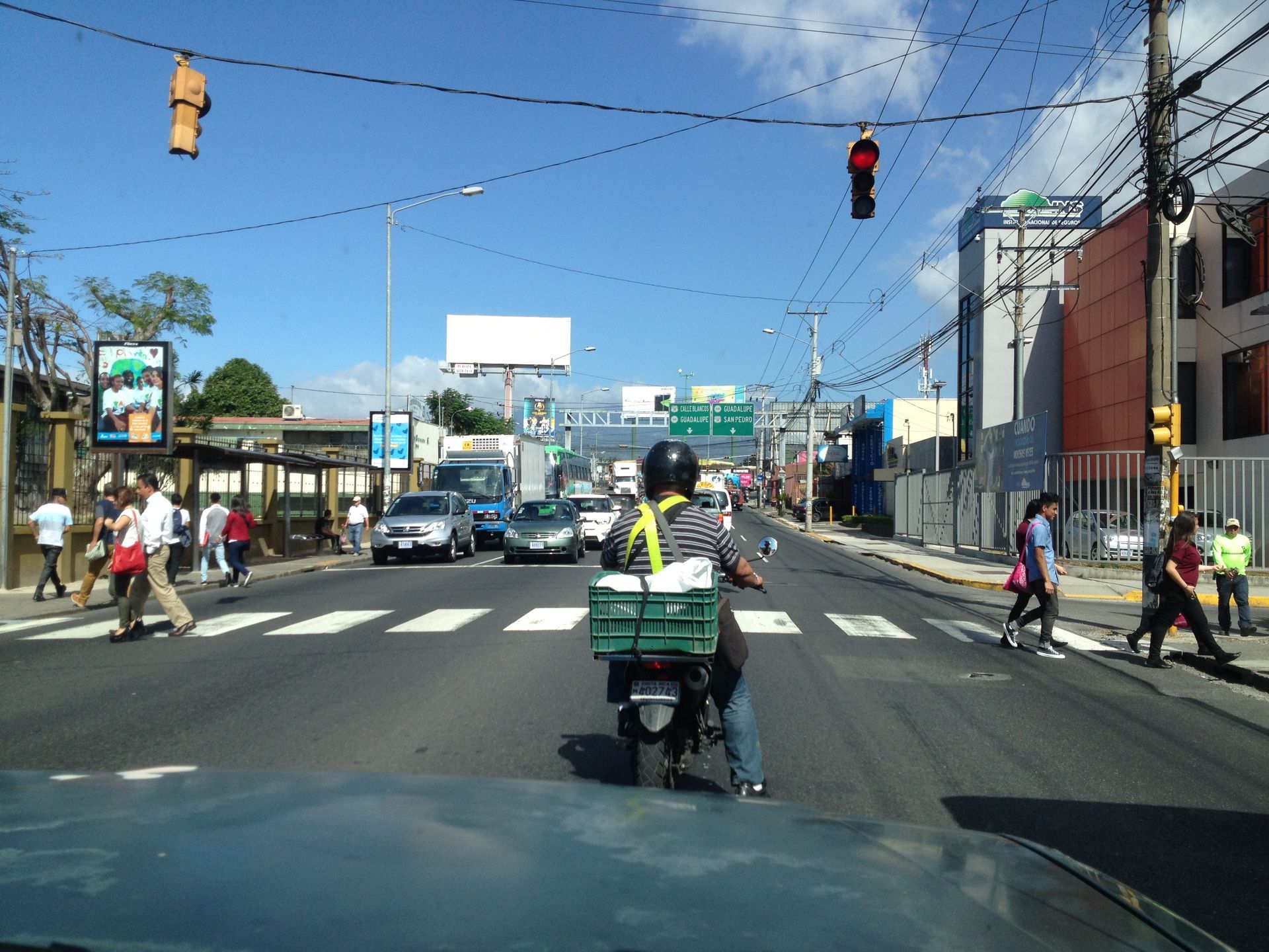 Motorcycle on road with traffic, pedestrians crossing crosswalk, buildings and blue sky.