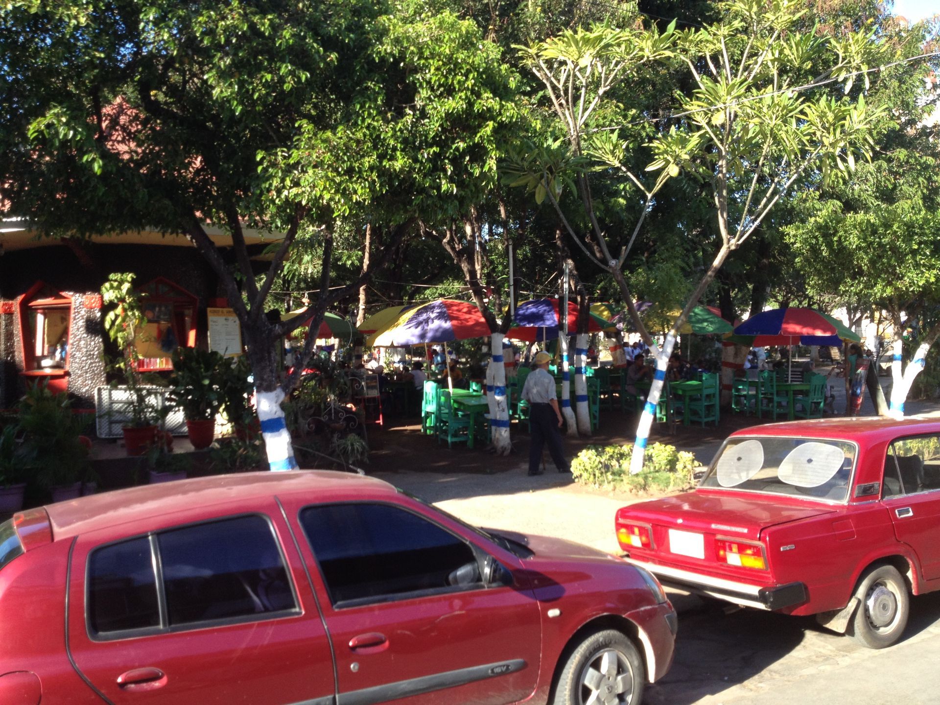 Street scene with parked red cars and a park area with tables, trees, and vendors under colorful umbrellas.