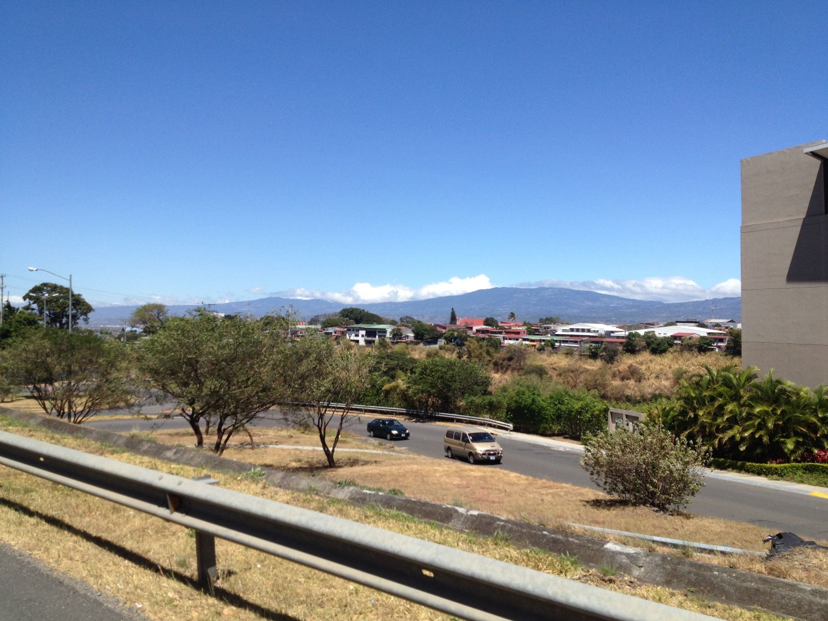 Road with cars, trees, buildings, and distant mountains under a blue sky.