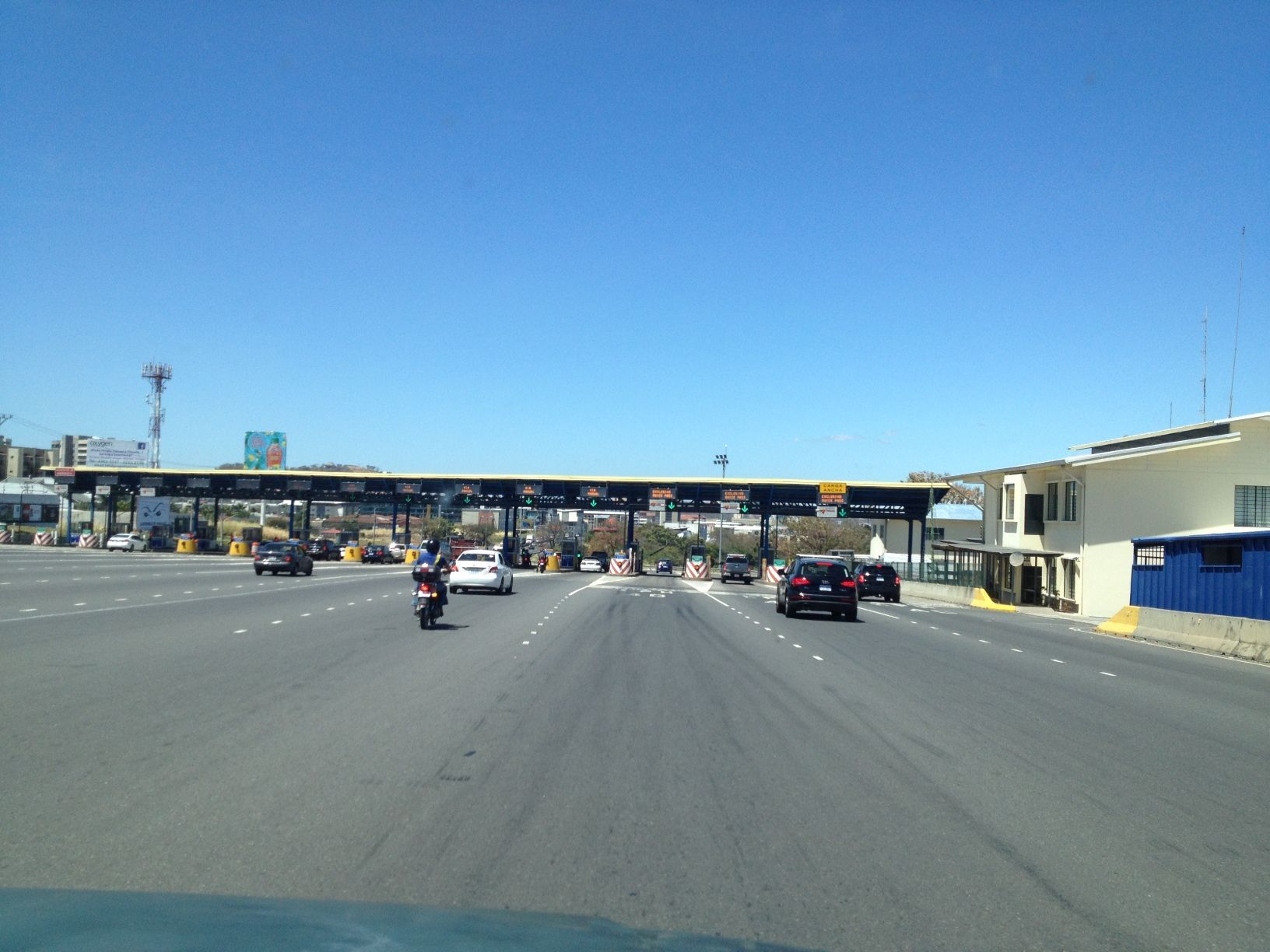 Cars and a motorcycle approach a toll booth under a clear blue sky.