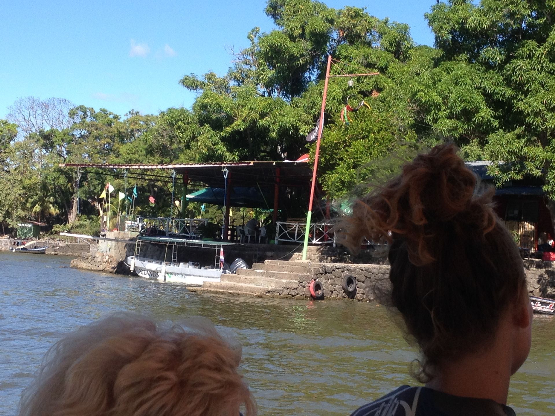 A person on a boat views a waterside restaurant under a canopy, surrounded by trees.