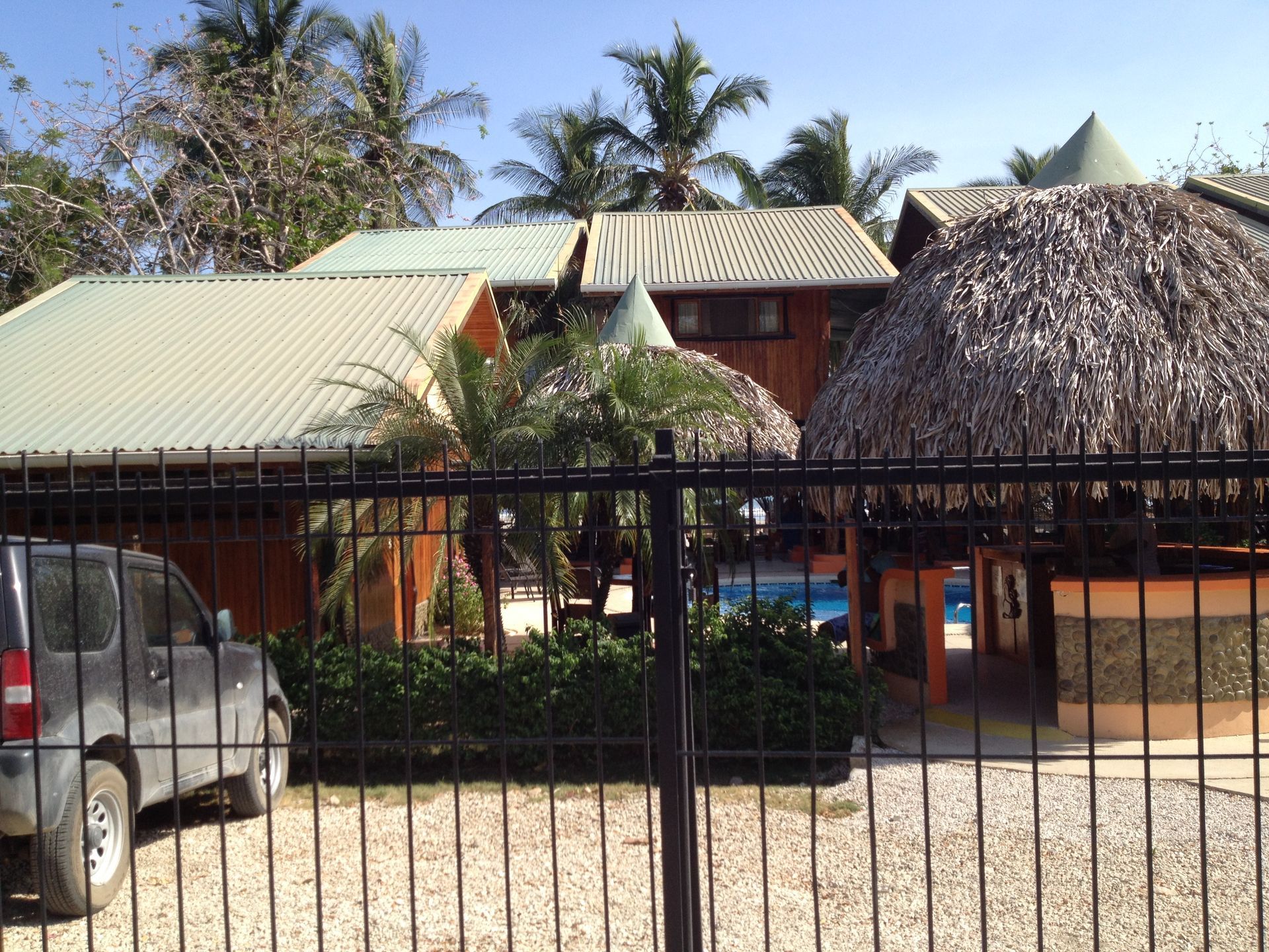 Wooden buildings with metal roofs and a thatched roof structure behind a black fence. A parked car is on the left.
