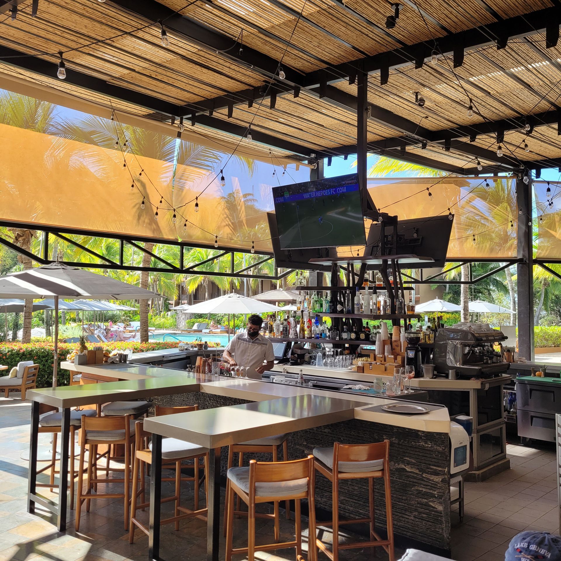 Outdoor bar with bartender, TVs, seating, and pool view.