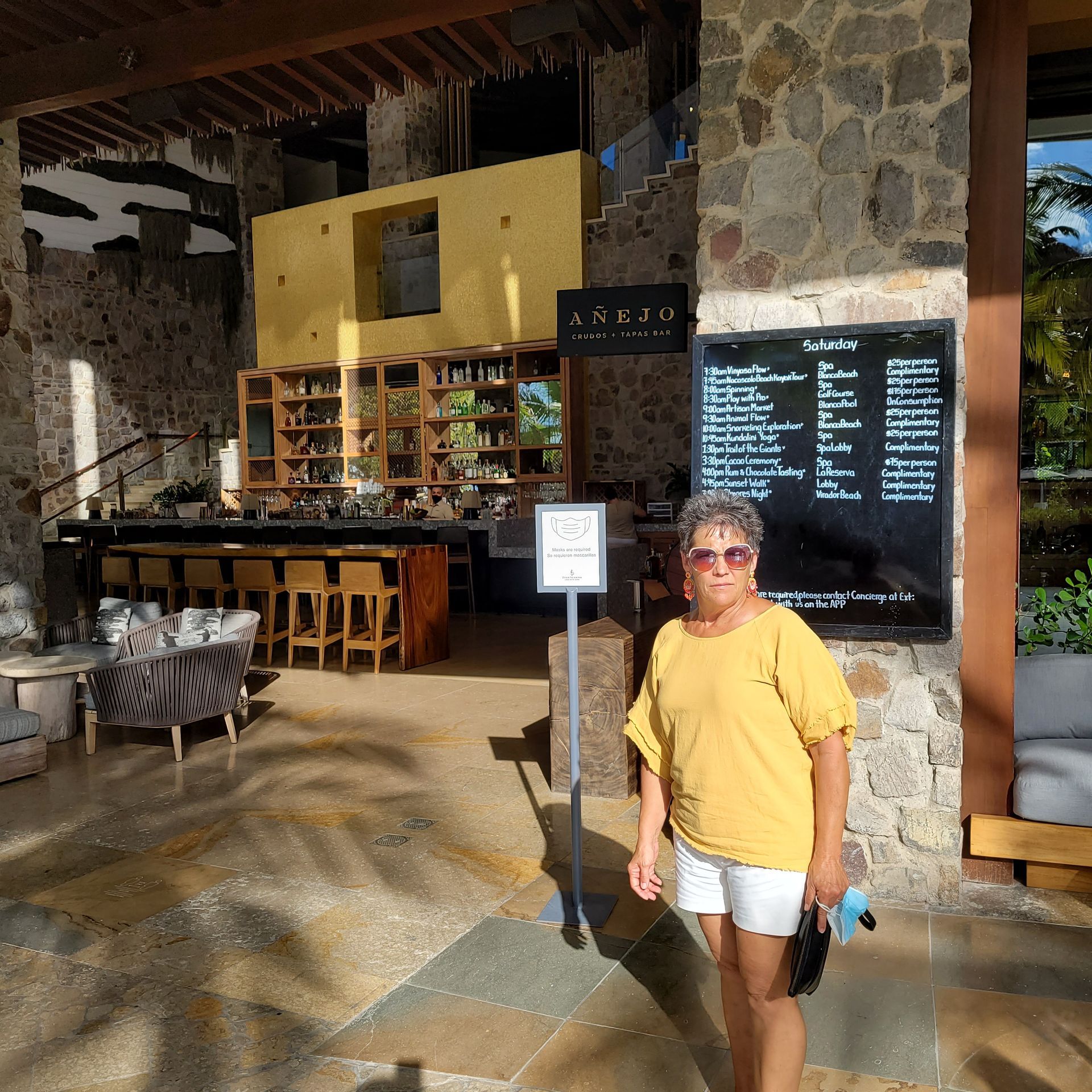 Woman in yellow shirt and white shorts stands outside a restaurant. Stone and wood building.