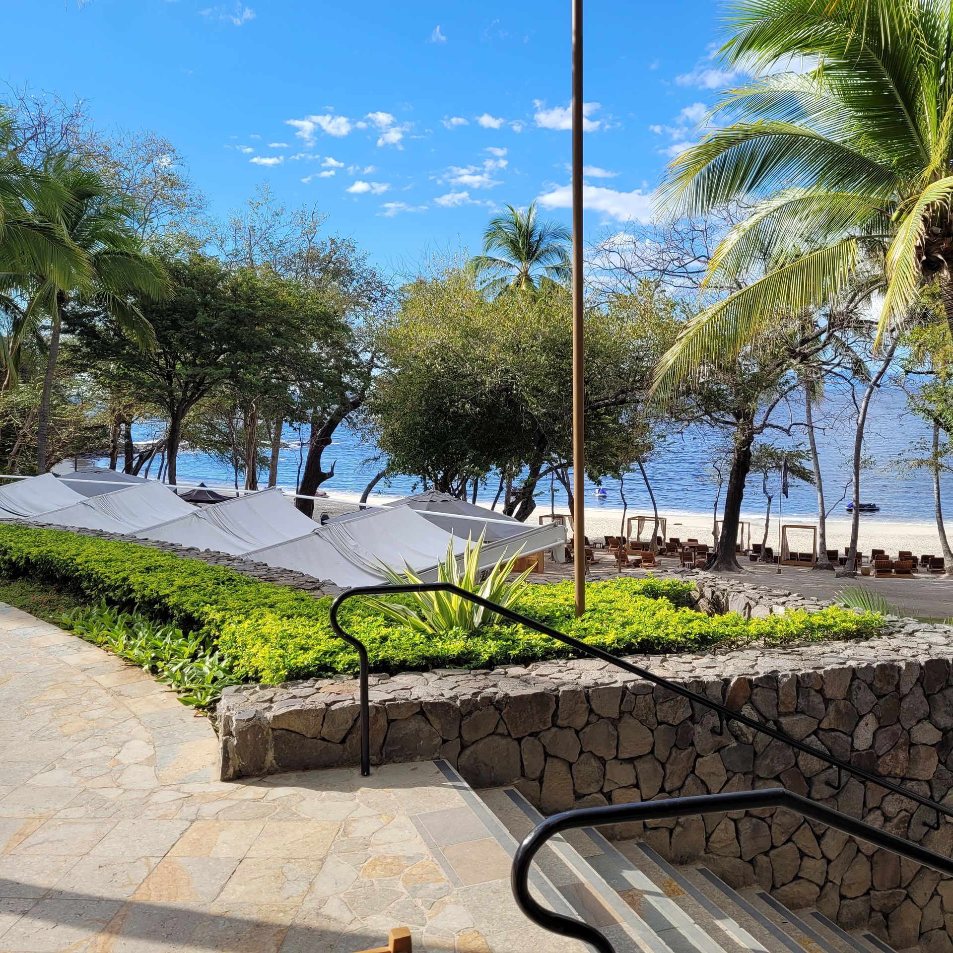 Stone steps lead to a beach with umbrellas, trees, and blue sky.