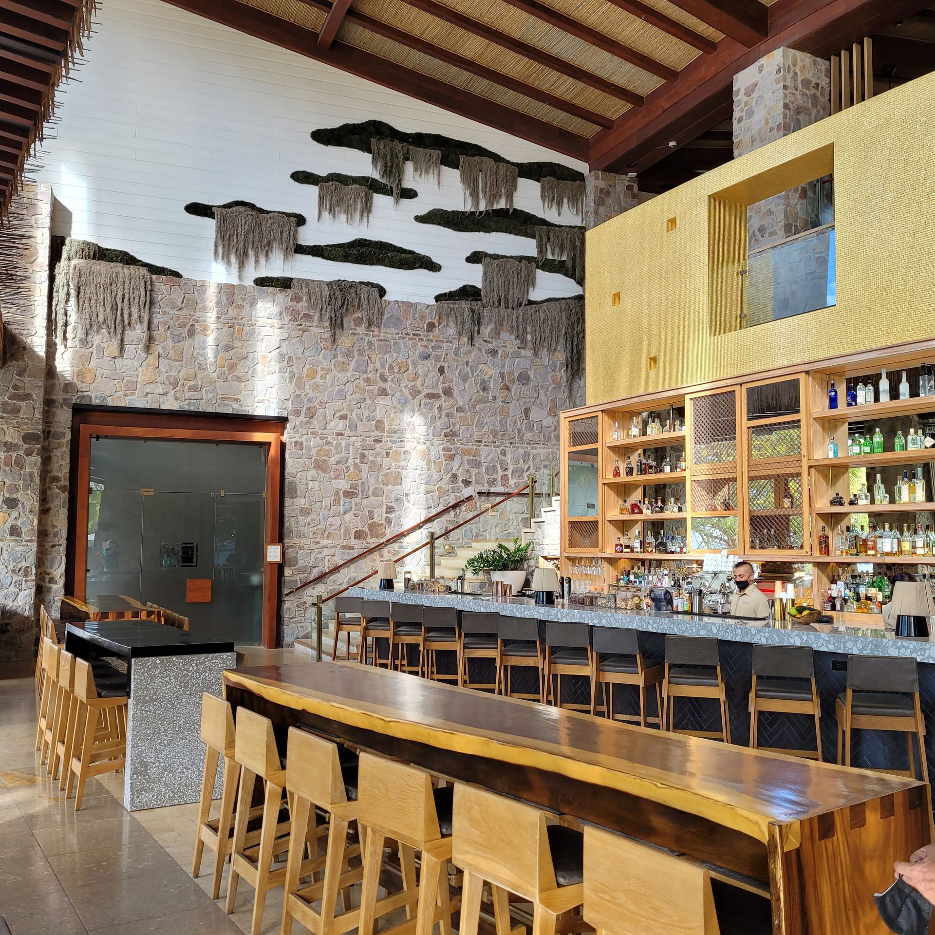 Bar interior: Stone walls, wood bar with stools, shelves of bottles, bartender, unique ceiling decor.