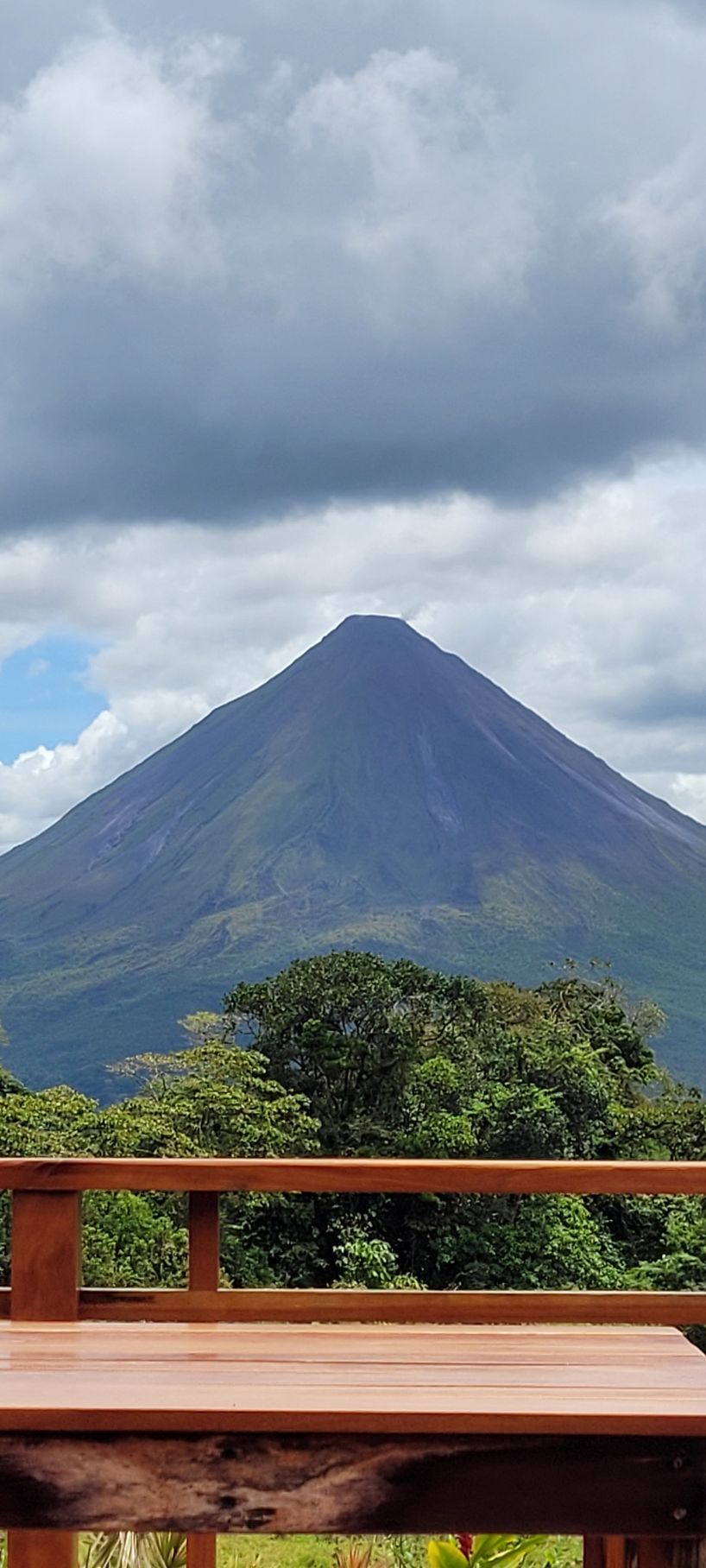 Arenal Volcano in Costa Rica framed by a wooden deck and lush greenery under a cloudy sky.