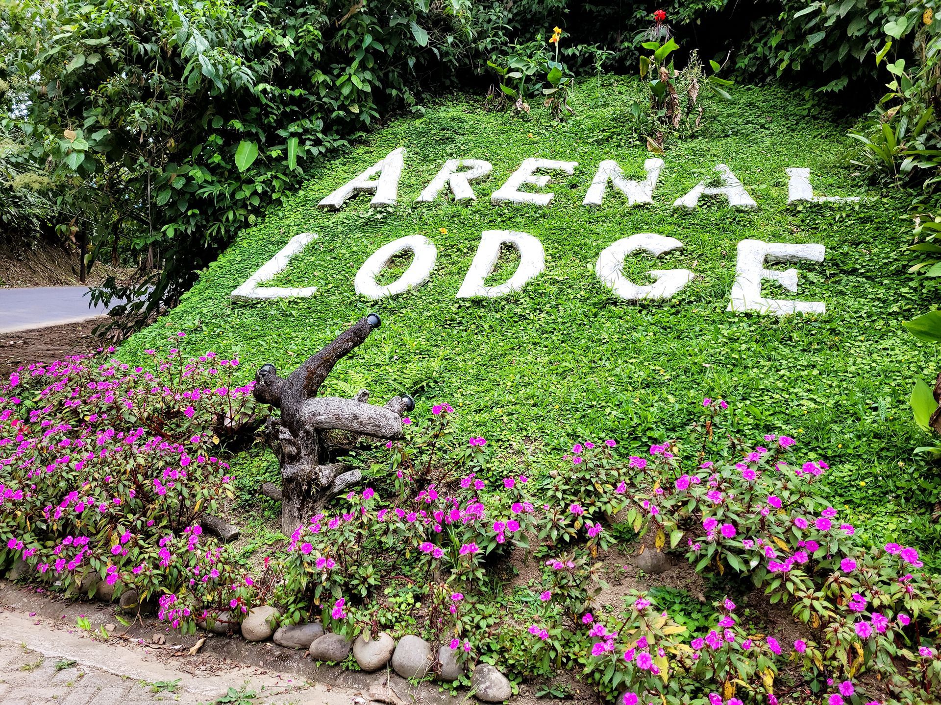 Arenal Lodge sign on a green, flowered hill. White letters, surrounded by purple blooms, with a sculpture in front.