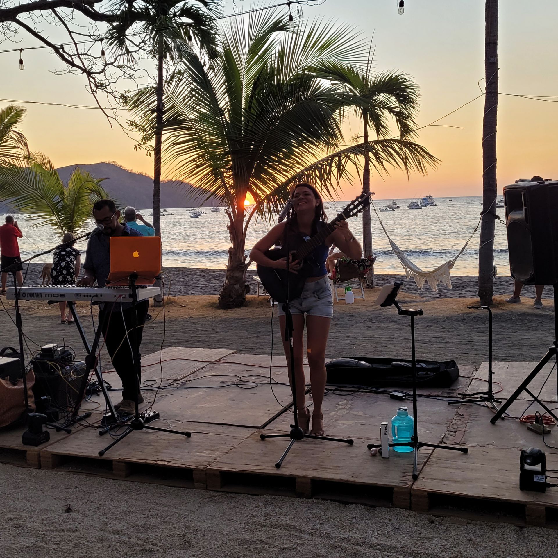 Musicians perform on a beach stage at sunset. A woman plays guitar, a man plays a keyboard; ocean in the background.