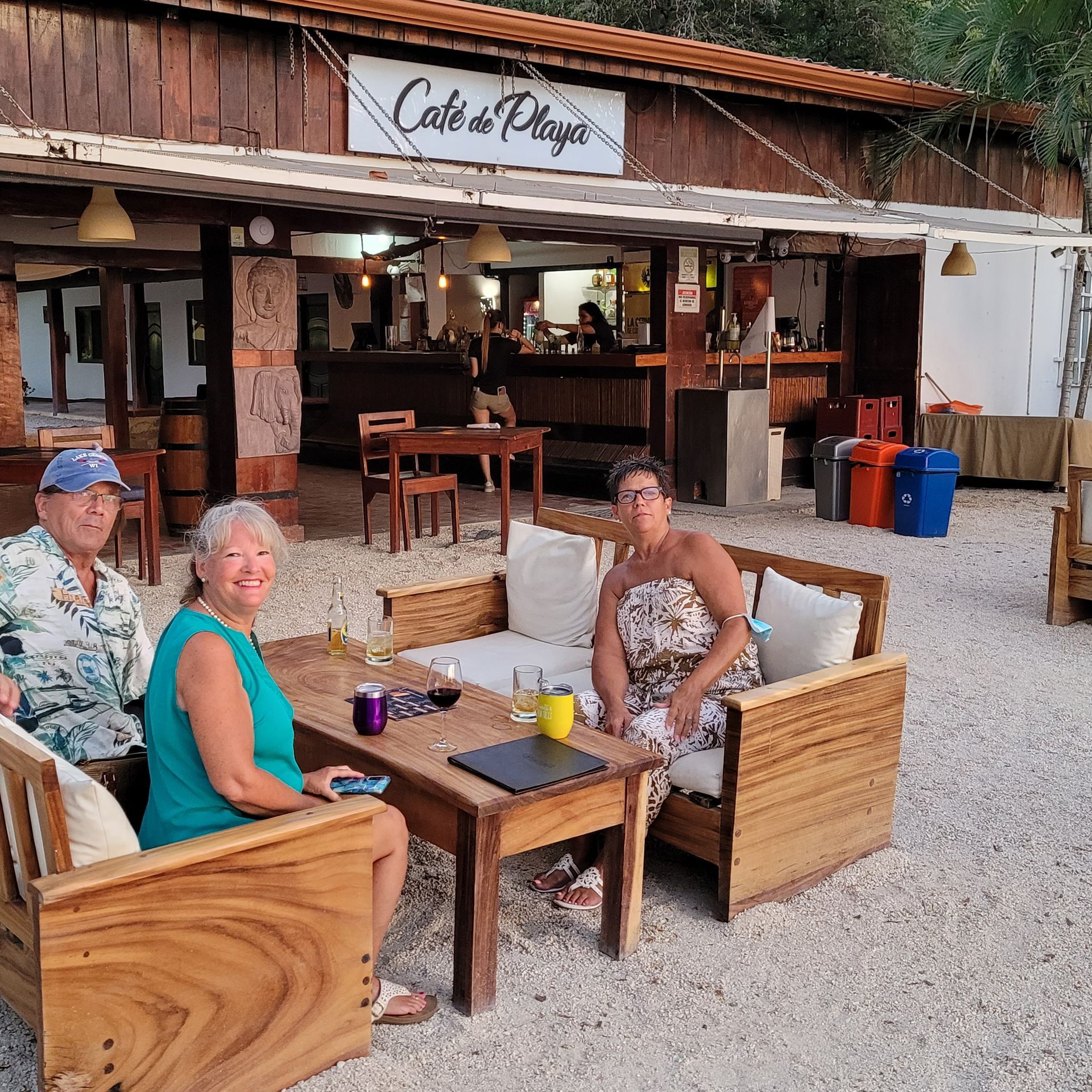 Three people seated on a wooden bench at Café de Playa.  Building with a bar in the background. Gravel ground.