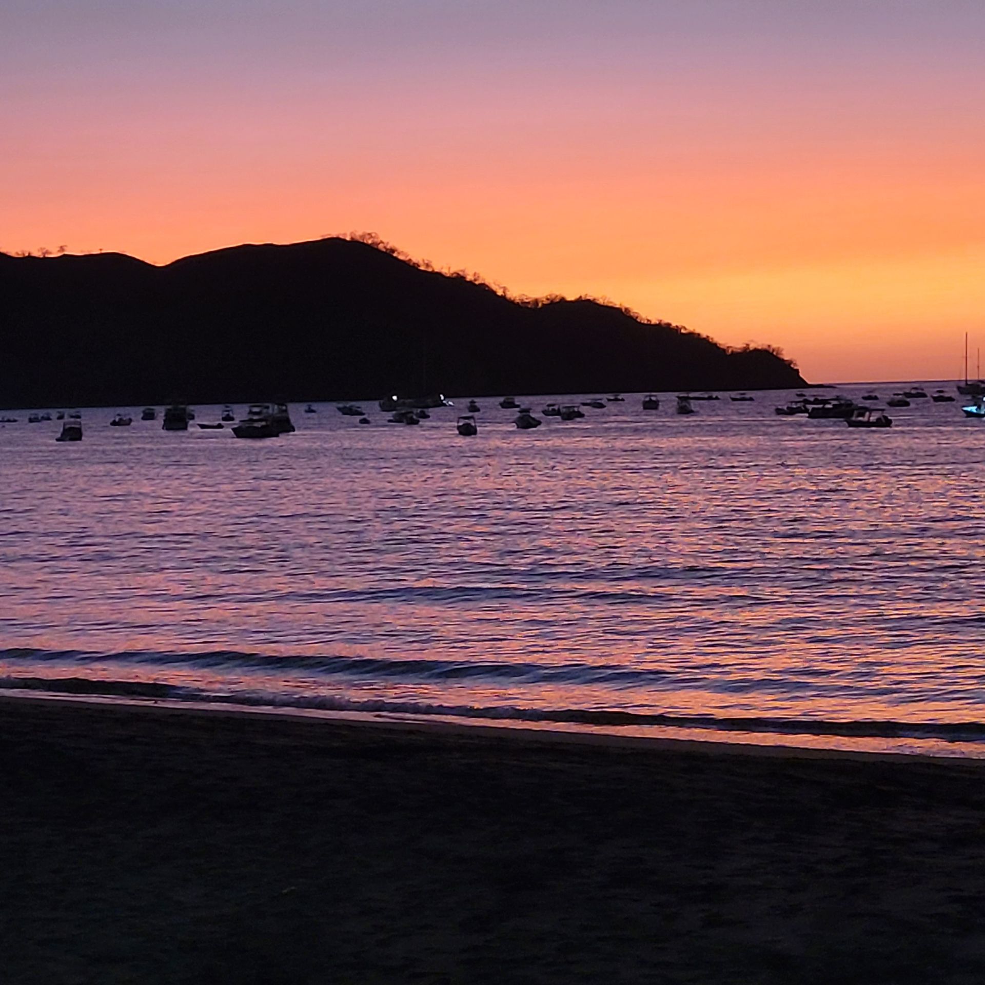Sunset over ocean with boats; orange and purple sky, dark silhouette of land.