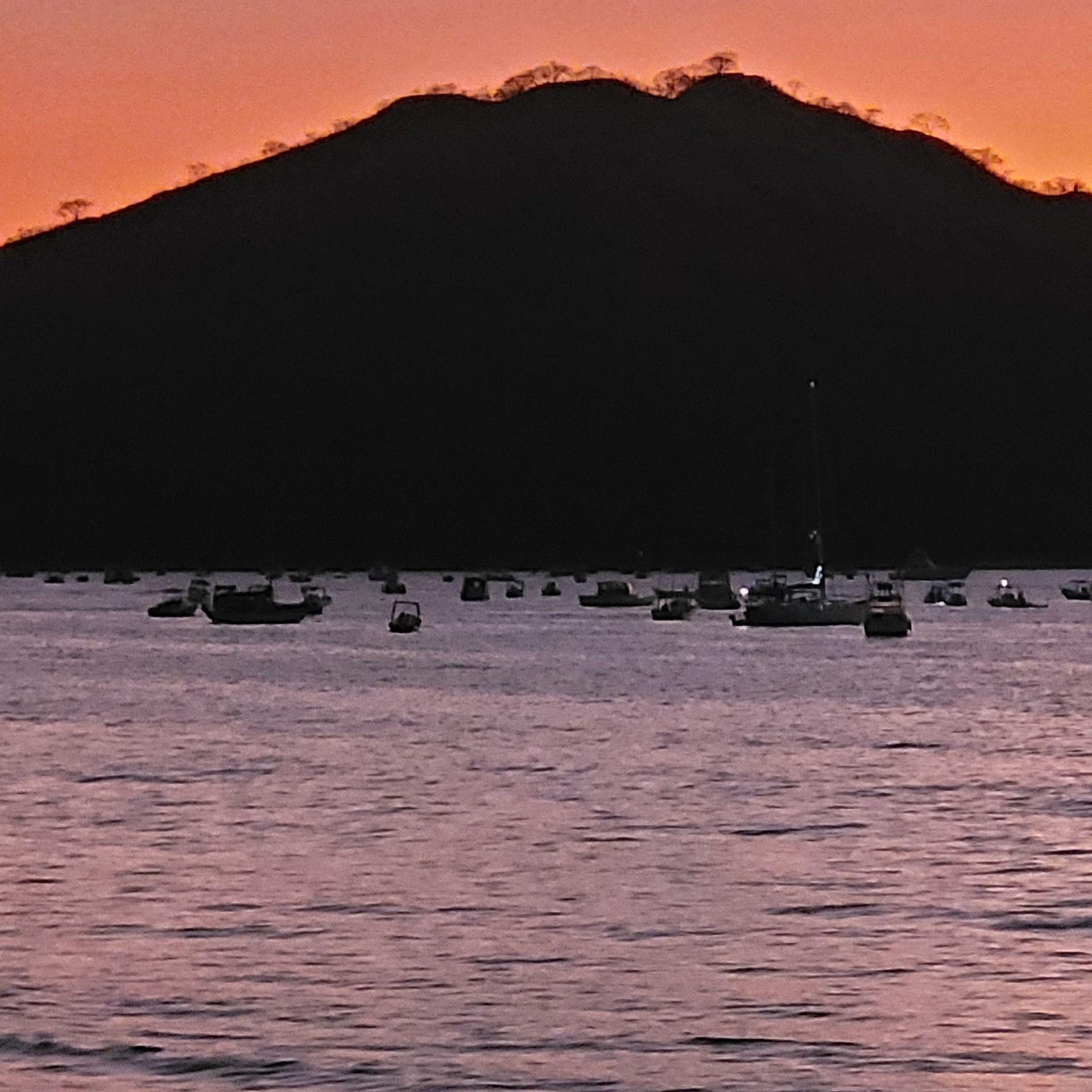 Boats on water in front of a silhouetted mountain at sunset with orange and purple hues.