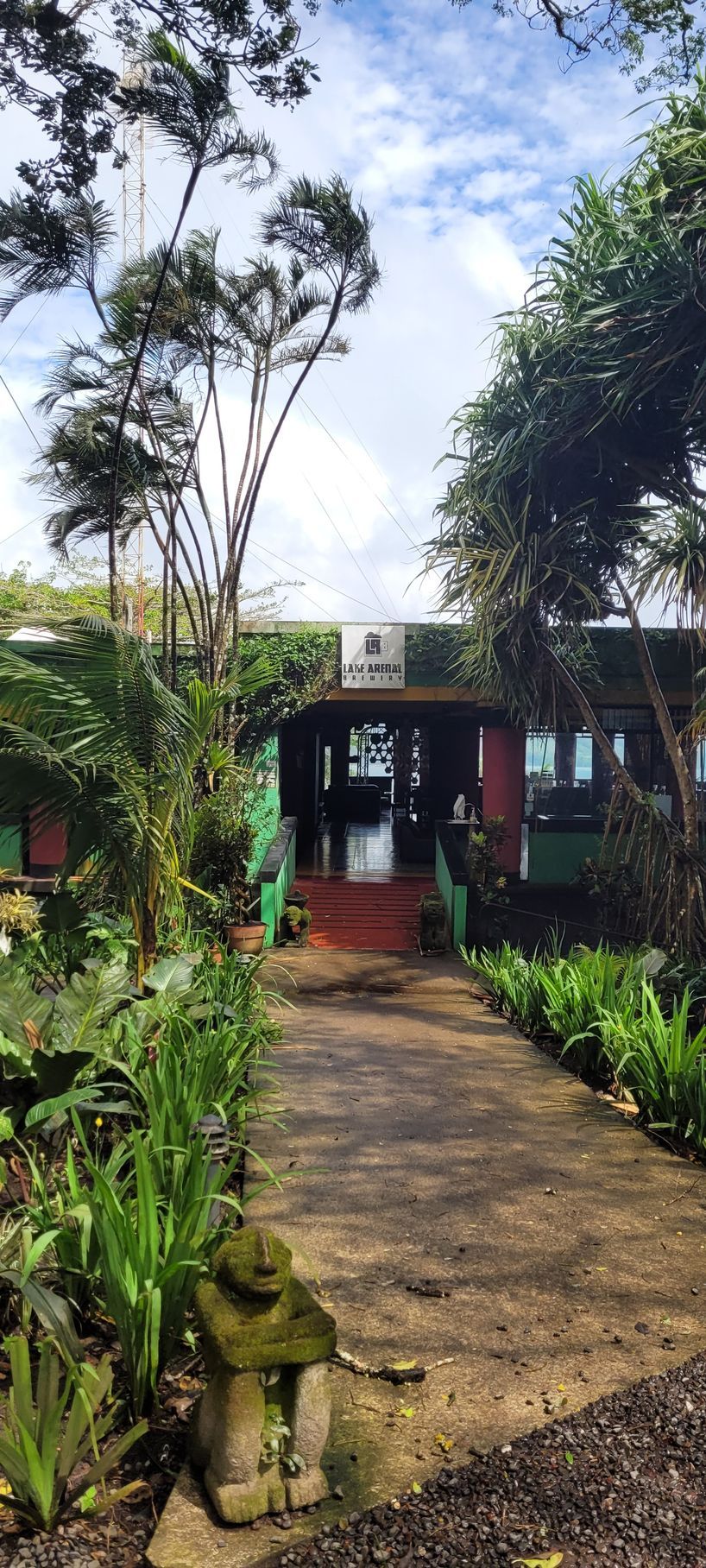 Pathway leads to a building with red steps, surrounded by lush green plants and trees under a cloudy sky.