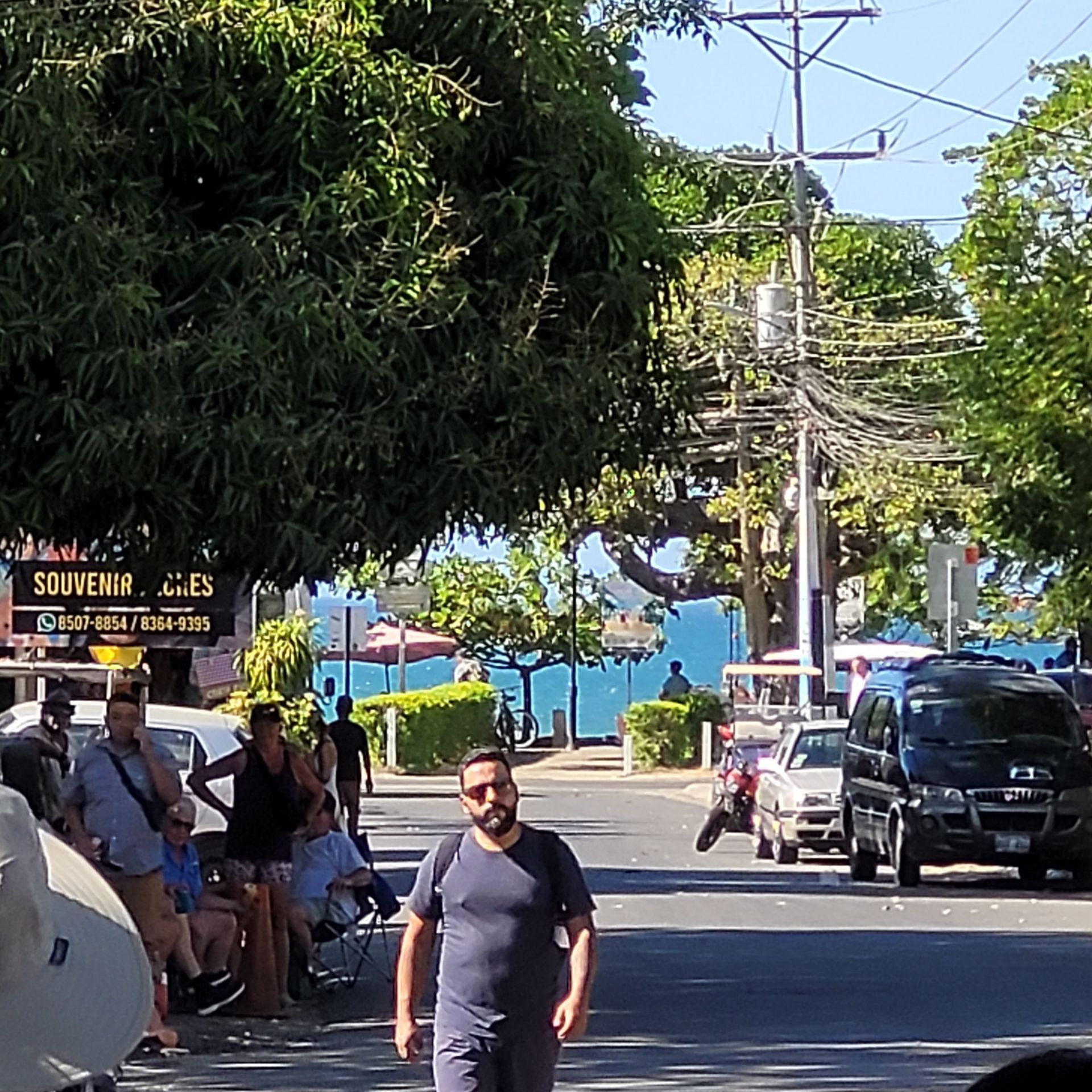 Street scene with people, cars, shops, and ocean view on a sunny day.