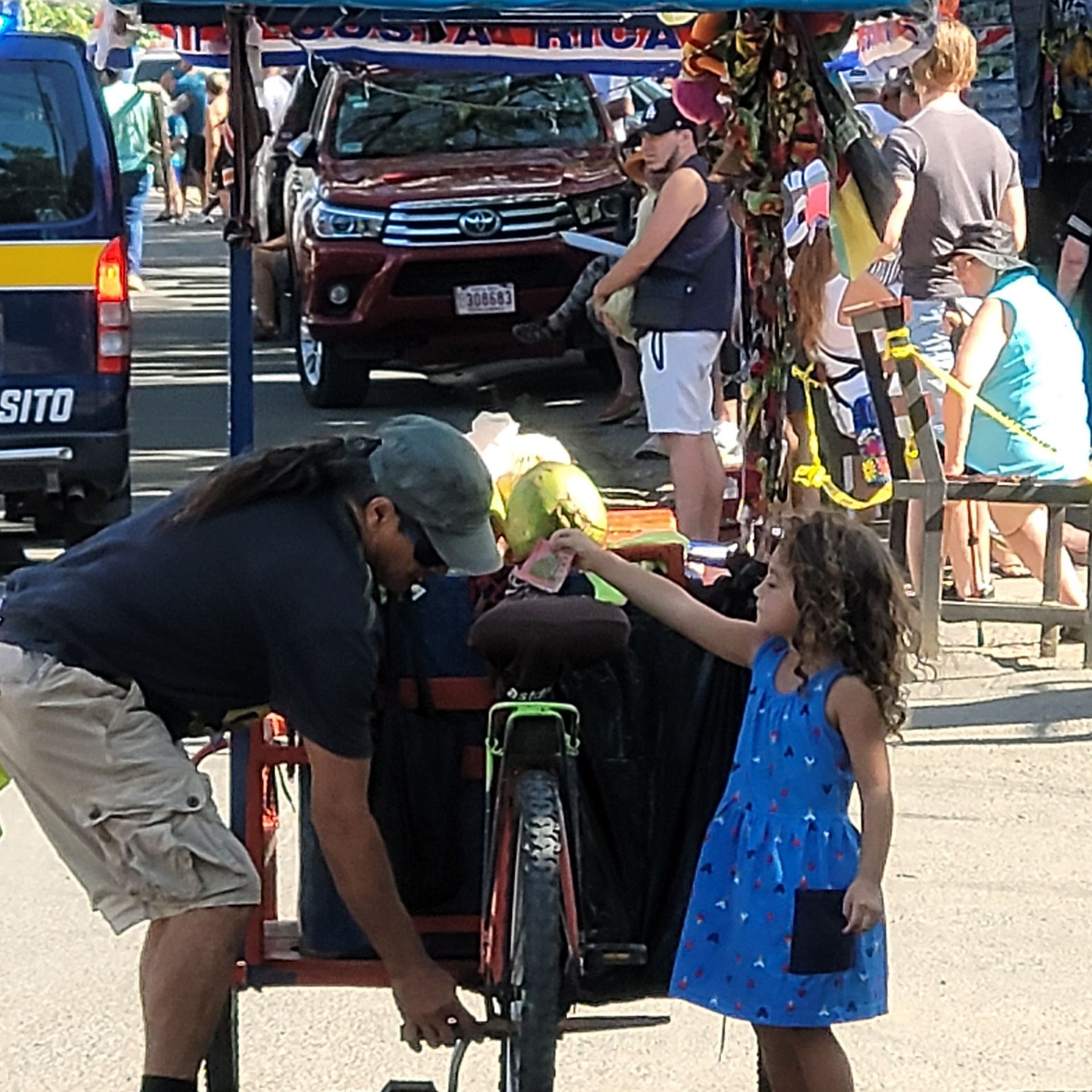 Man fixing a bike as a young child reaches toward him, market in the background with cars and people.