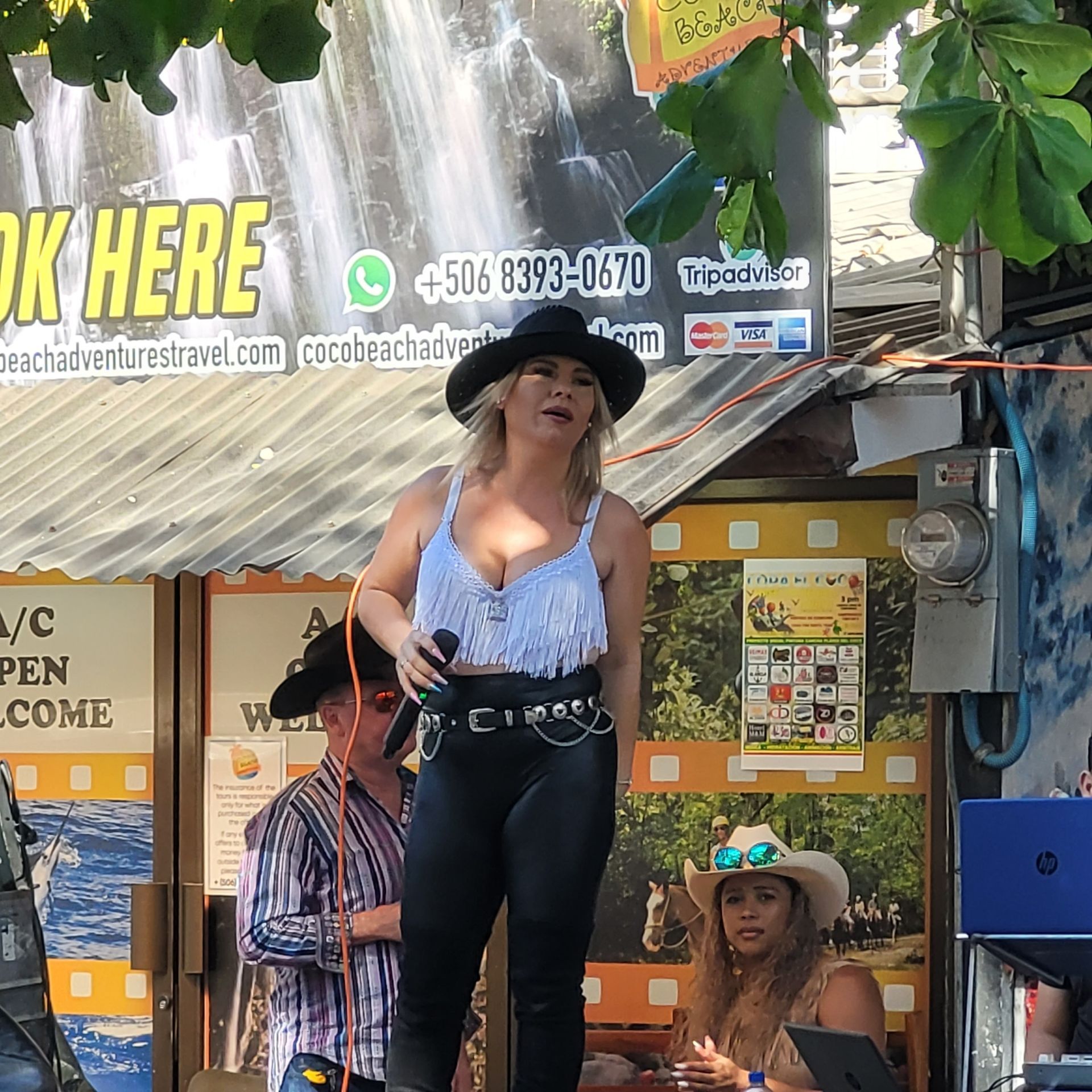 Woman singing, wearing hat and fringe top, performing outdoors at a travel agency.