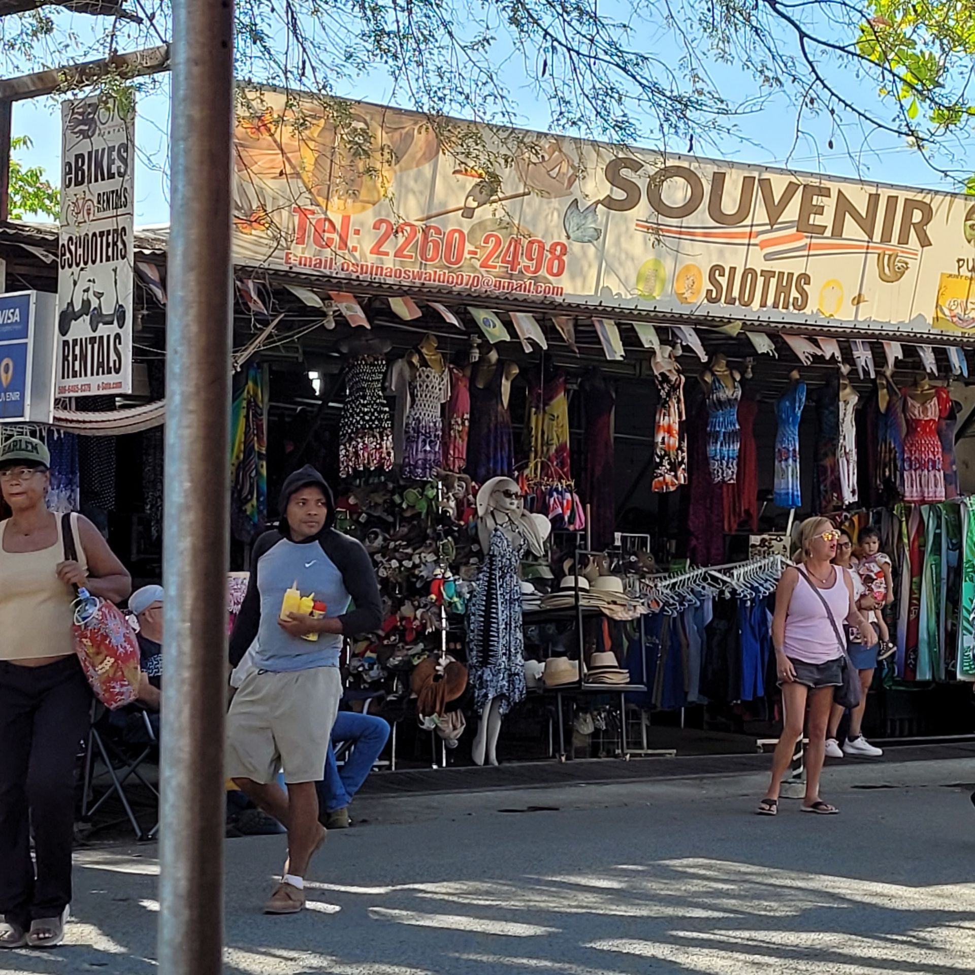Street vendor shop with clothing and souvenirs; people walking by on a sunny day.