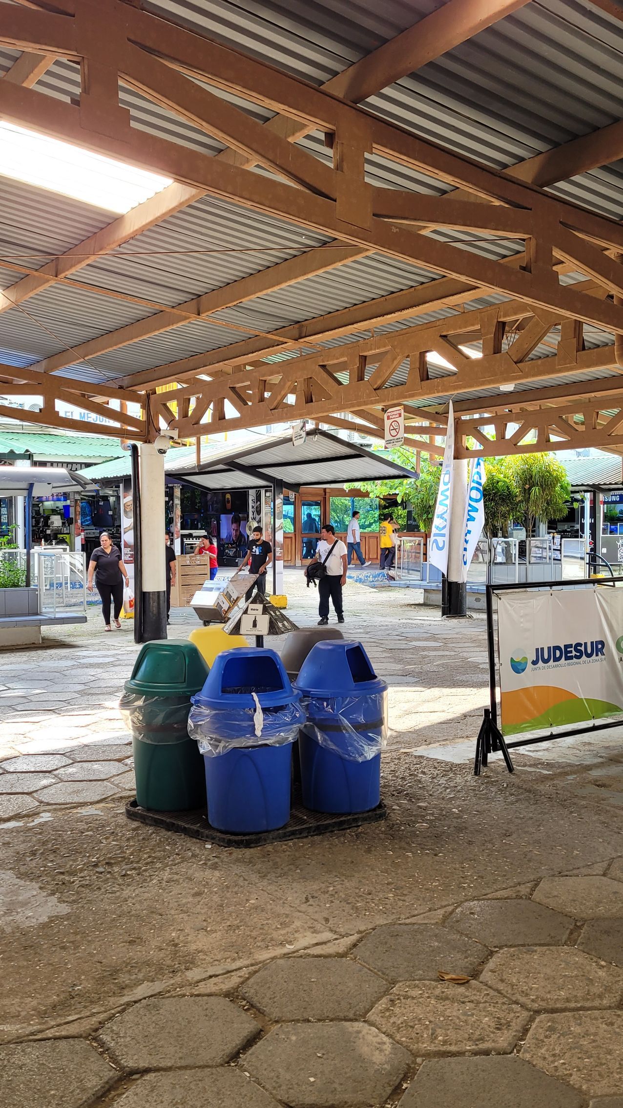 Three recycling bins under a wooden-beamed awning. People in the background near shops.