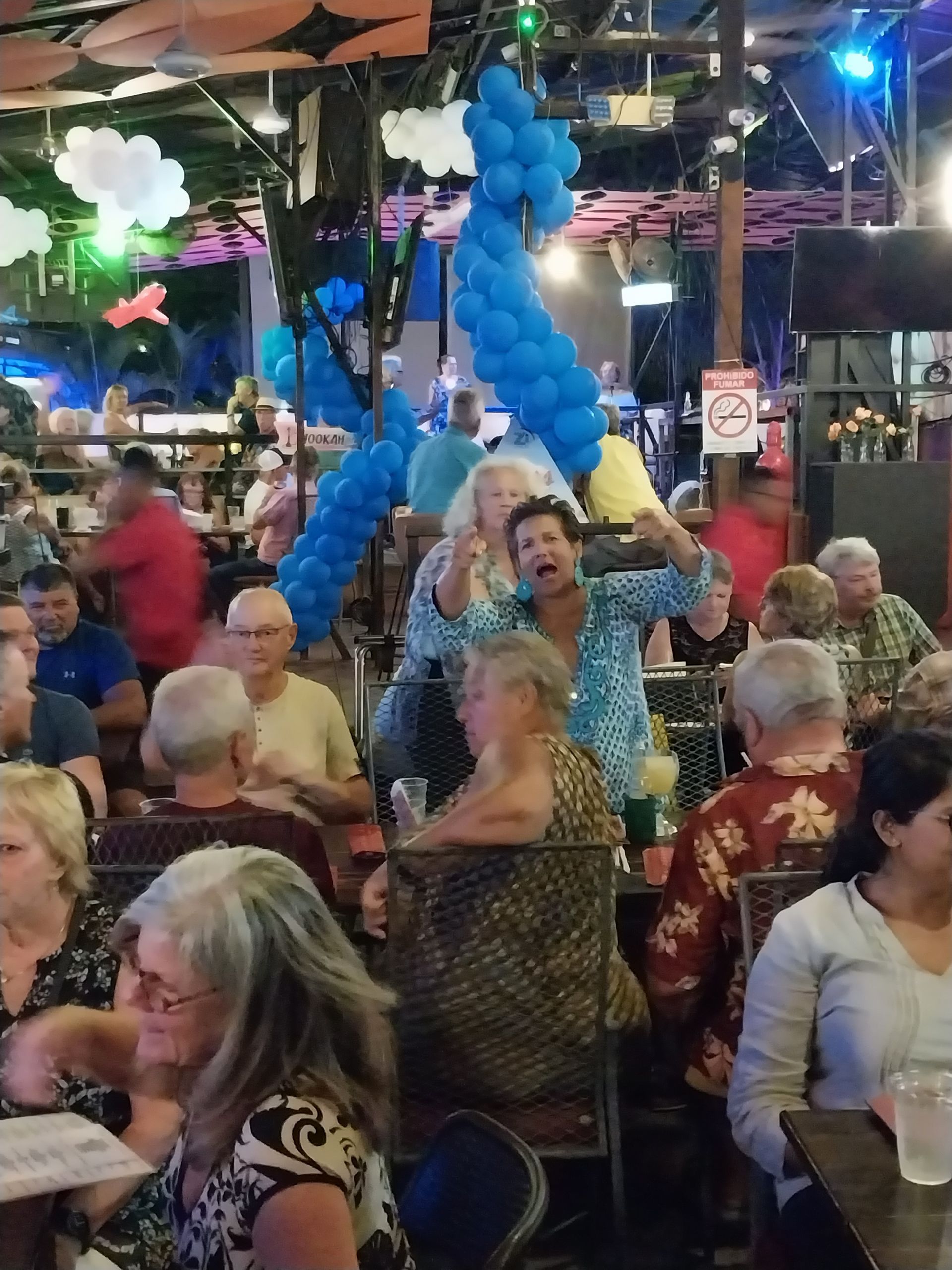 A crowded restaurant with people and blue balloon decorations. A woman is enthusiastically posing in the center.