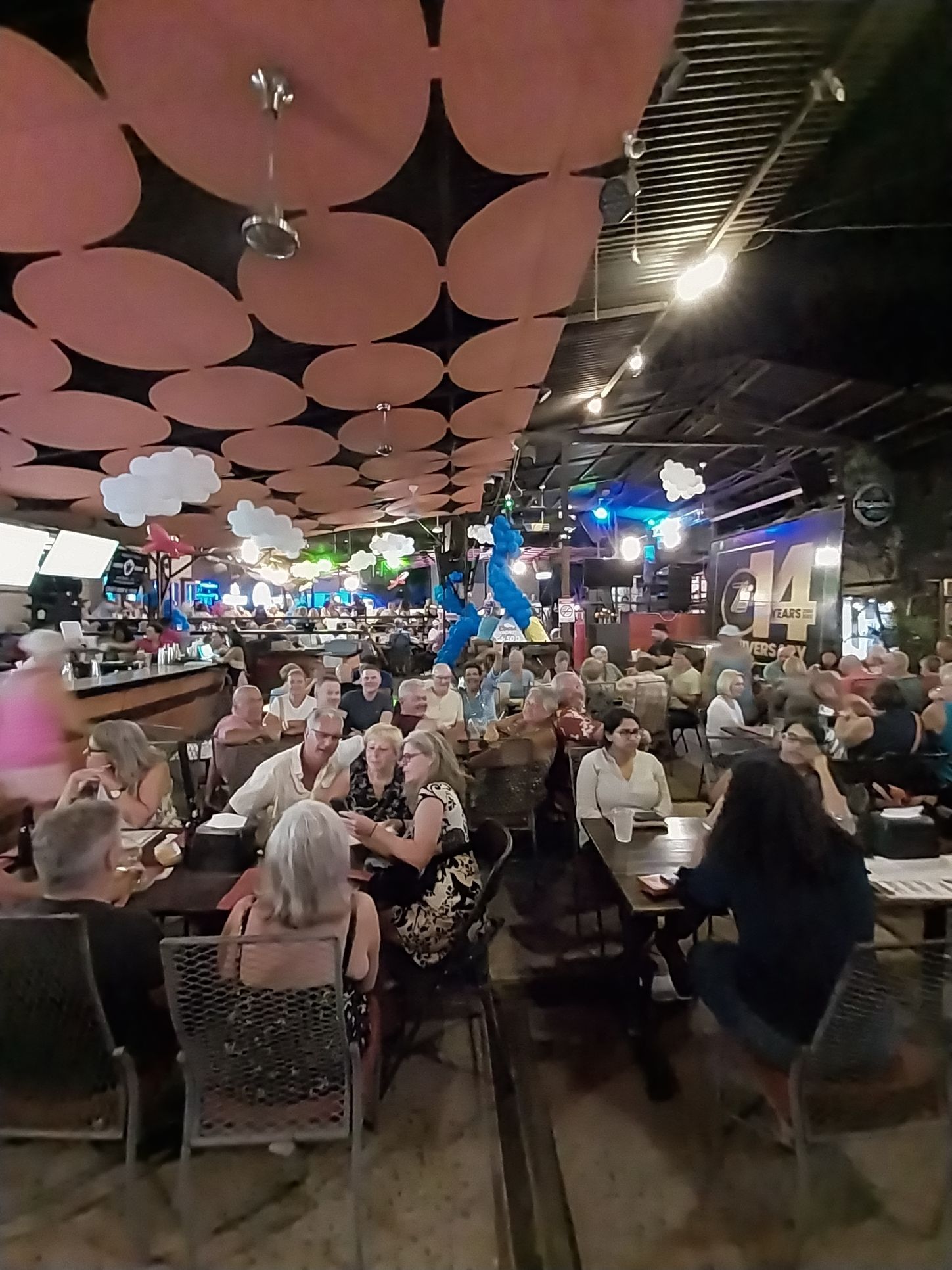 A crowded outdoor bar/restaurant with patrons seated at tables. Overhead, pink and black decor.