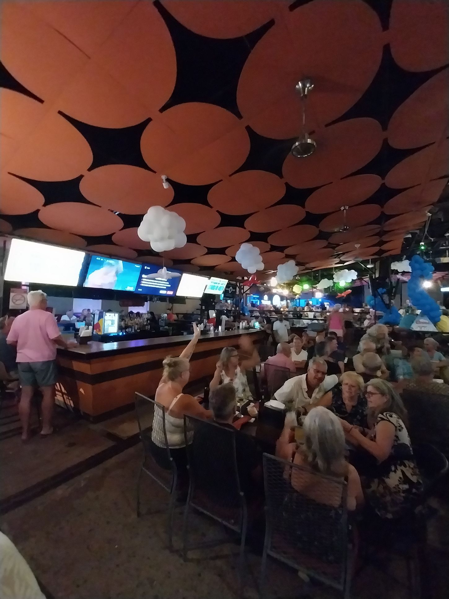 Interior of a bar, people seated at tables, watching TVs above the bar. Orange and black ceiling decor.