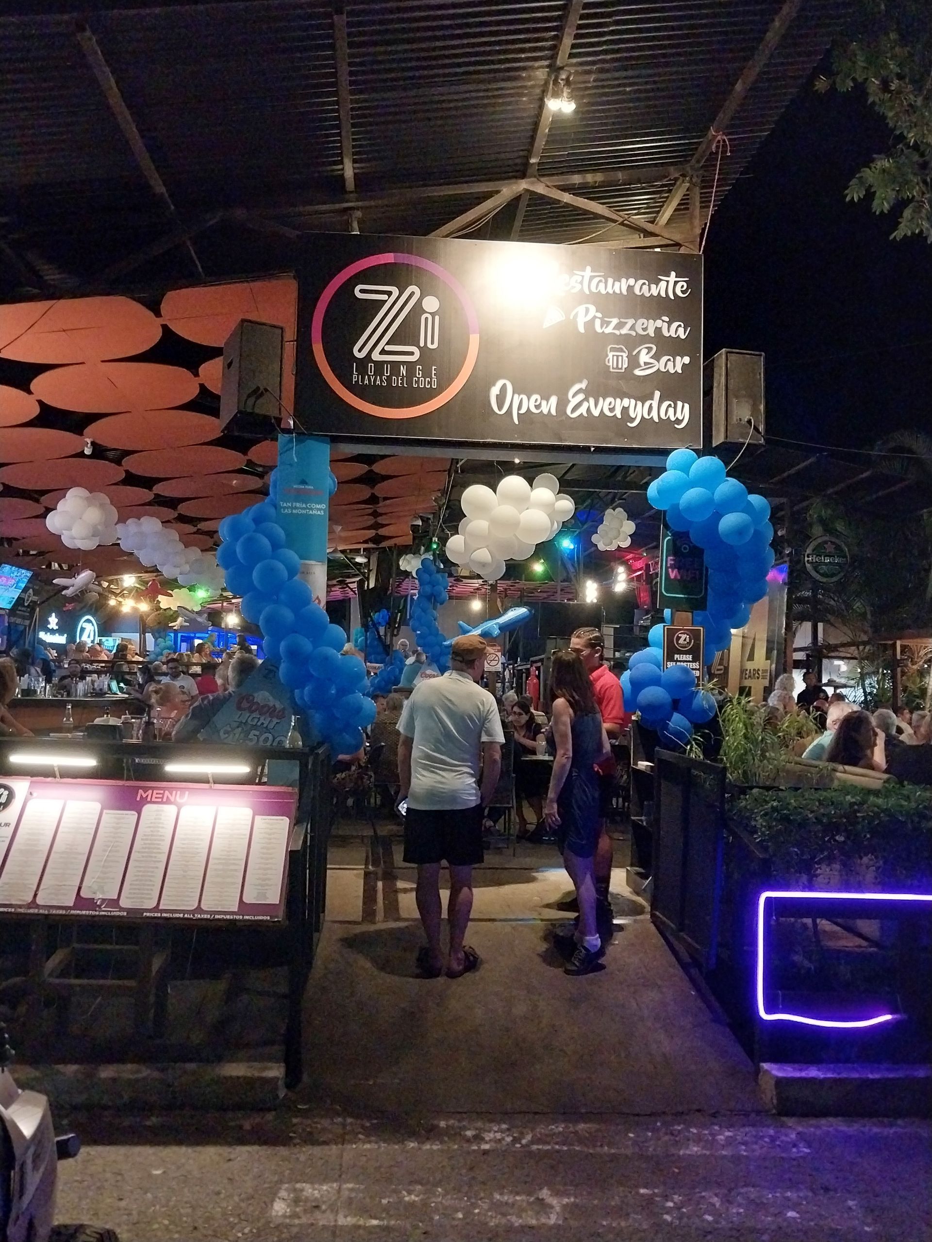 Entrance to Zi Restaurant & Pizzeria, decorated with balloons, people walking in, outdoor setting at night.