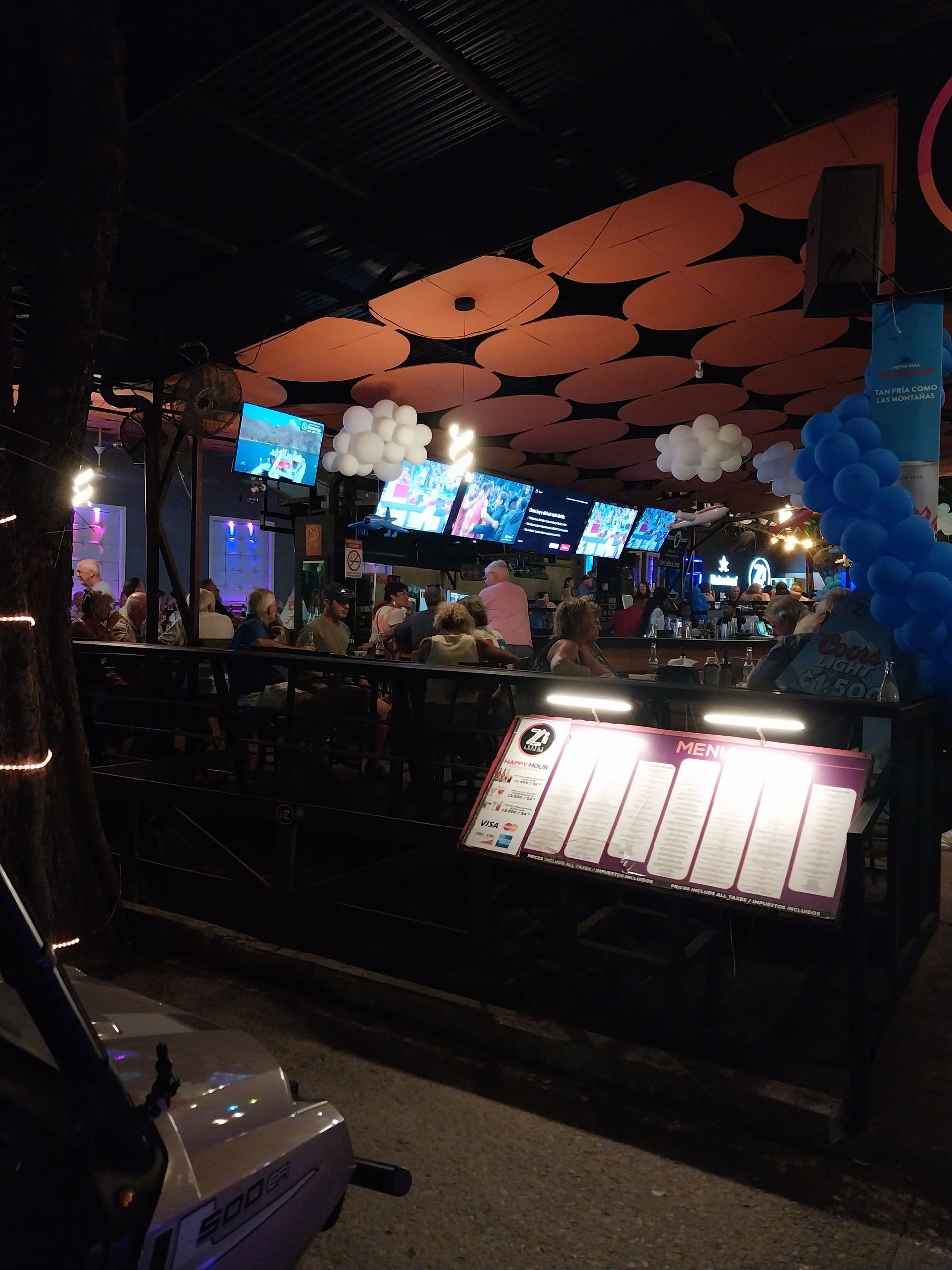 Bar interior with patrons seated at a long counter, multiple TVs, decorative ceiling, and menu stand.