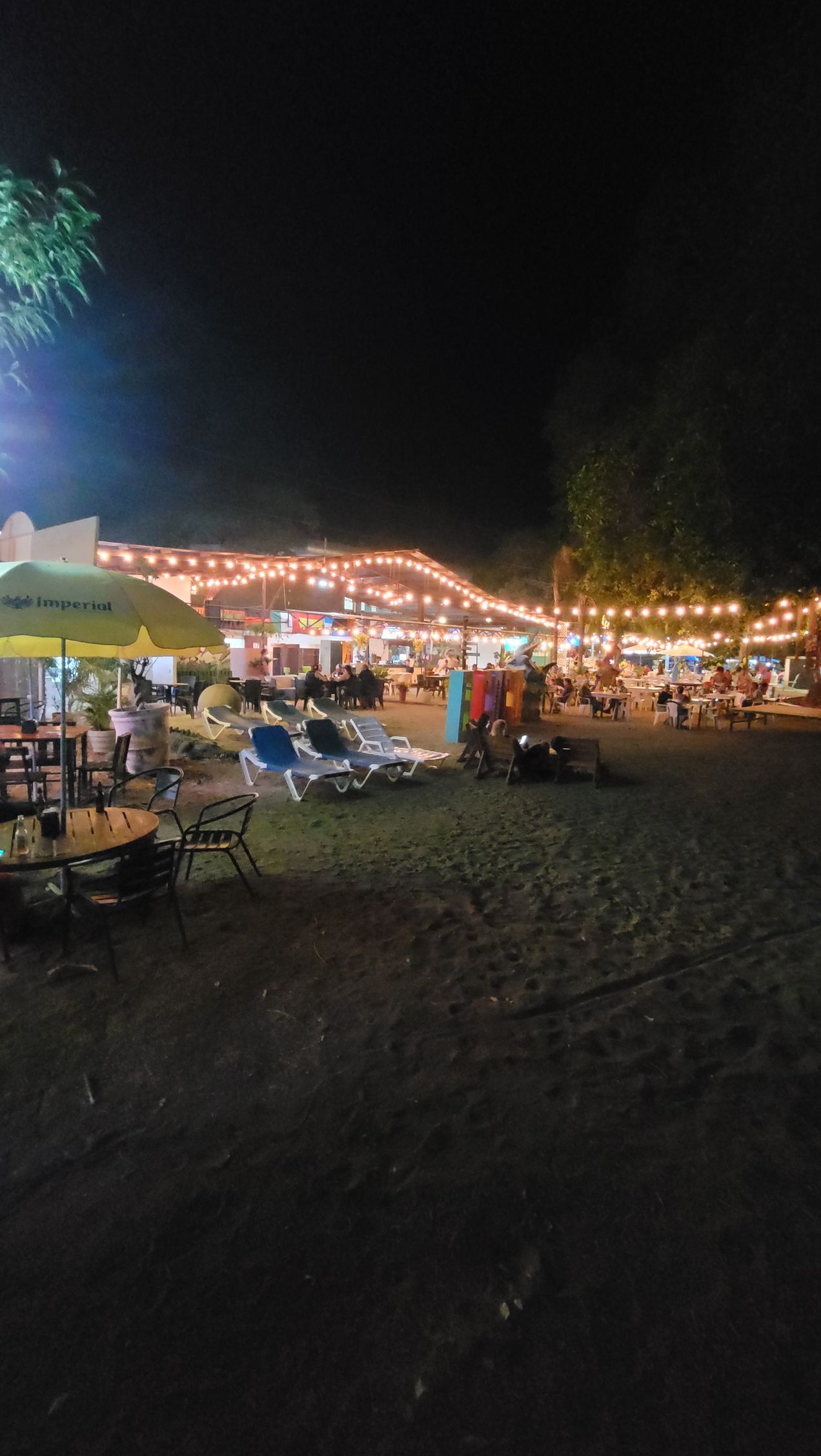 Beachside scene at night, lit with string lights. Tables, chairs, and people gathered.
