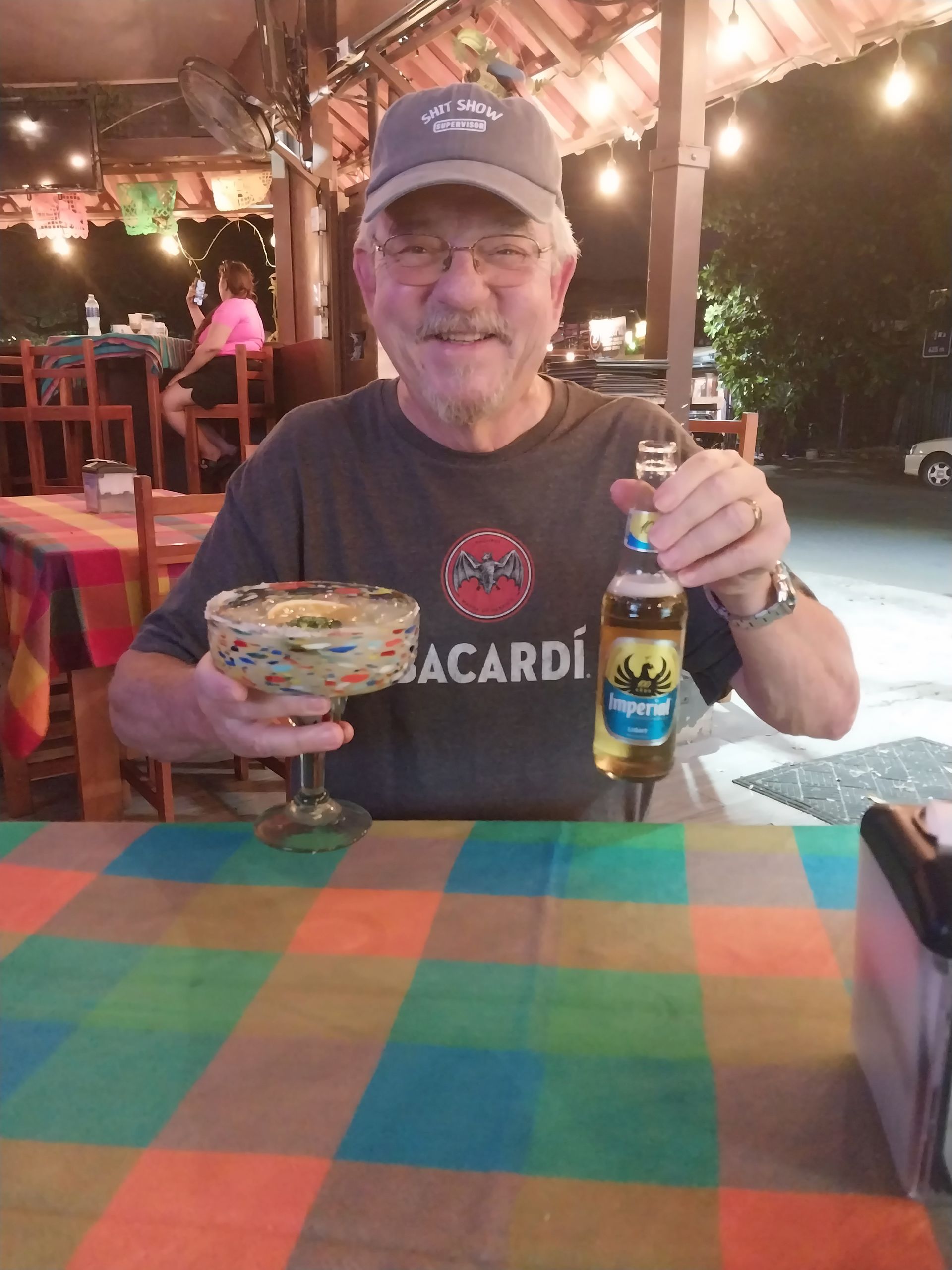 Man smiling, holding a margarita and beer bottle at an outdoor restaurant table.