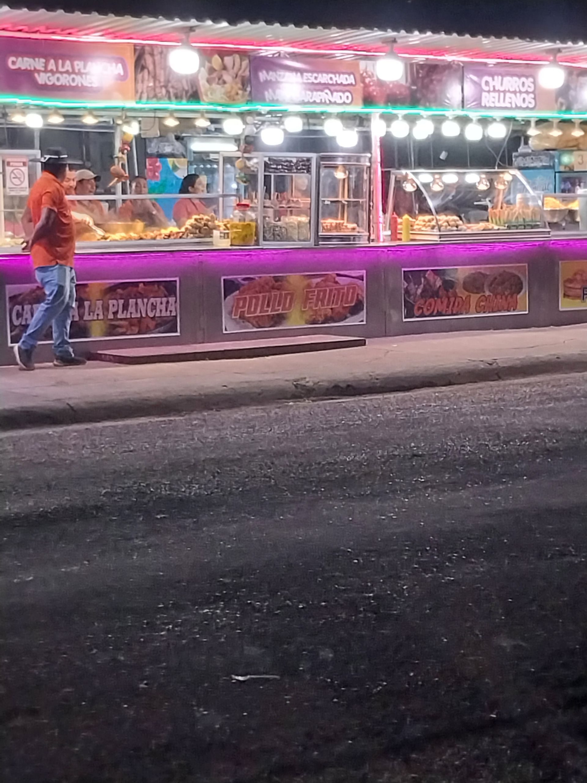 Food stand at night, displaying food options. A person walks past, lit by colorful lights.