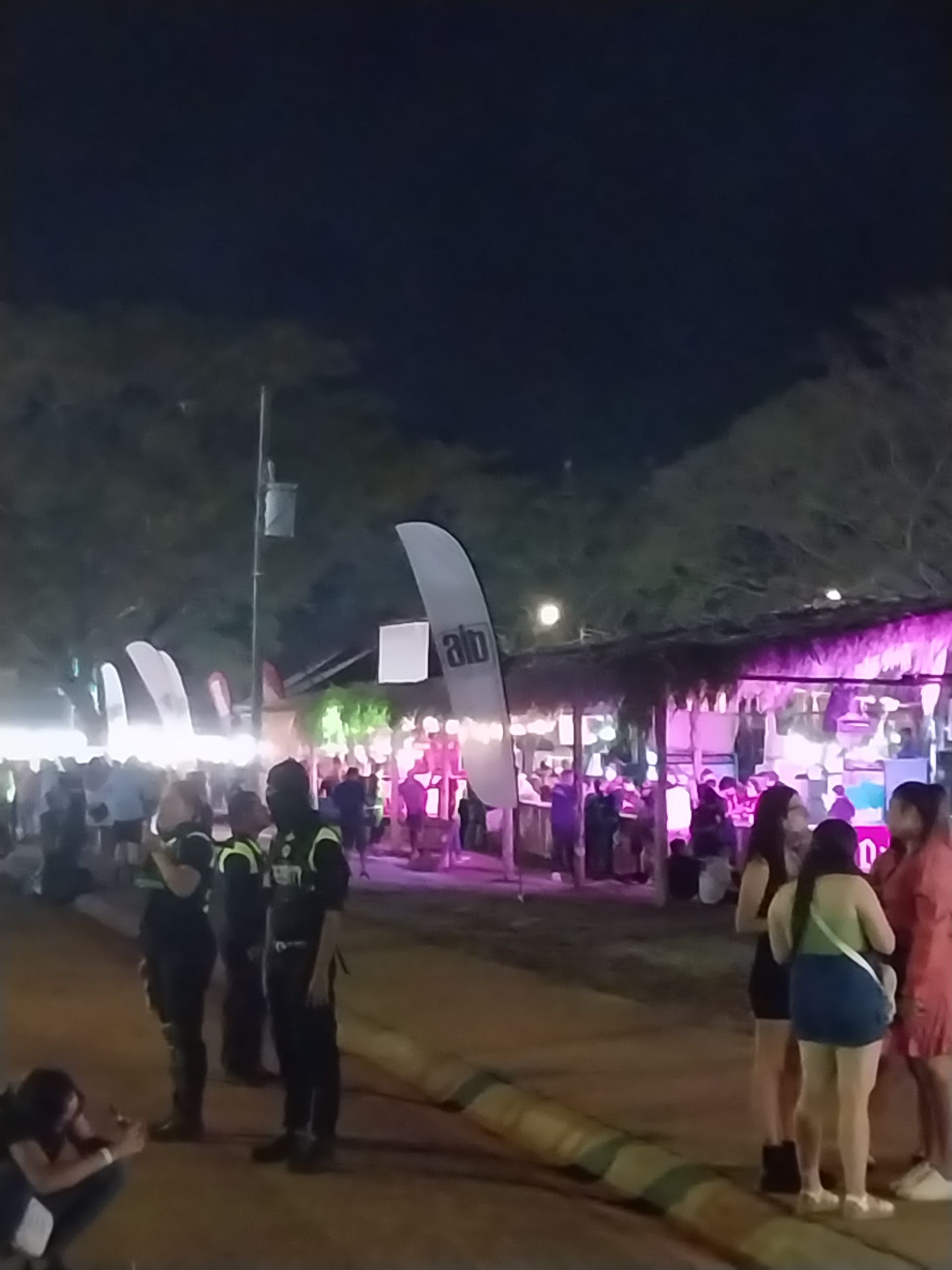 Nighttime outdoor event with a crowd. Stage lit in pink, people standing, security visible, Alo flags.