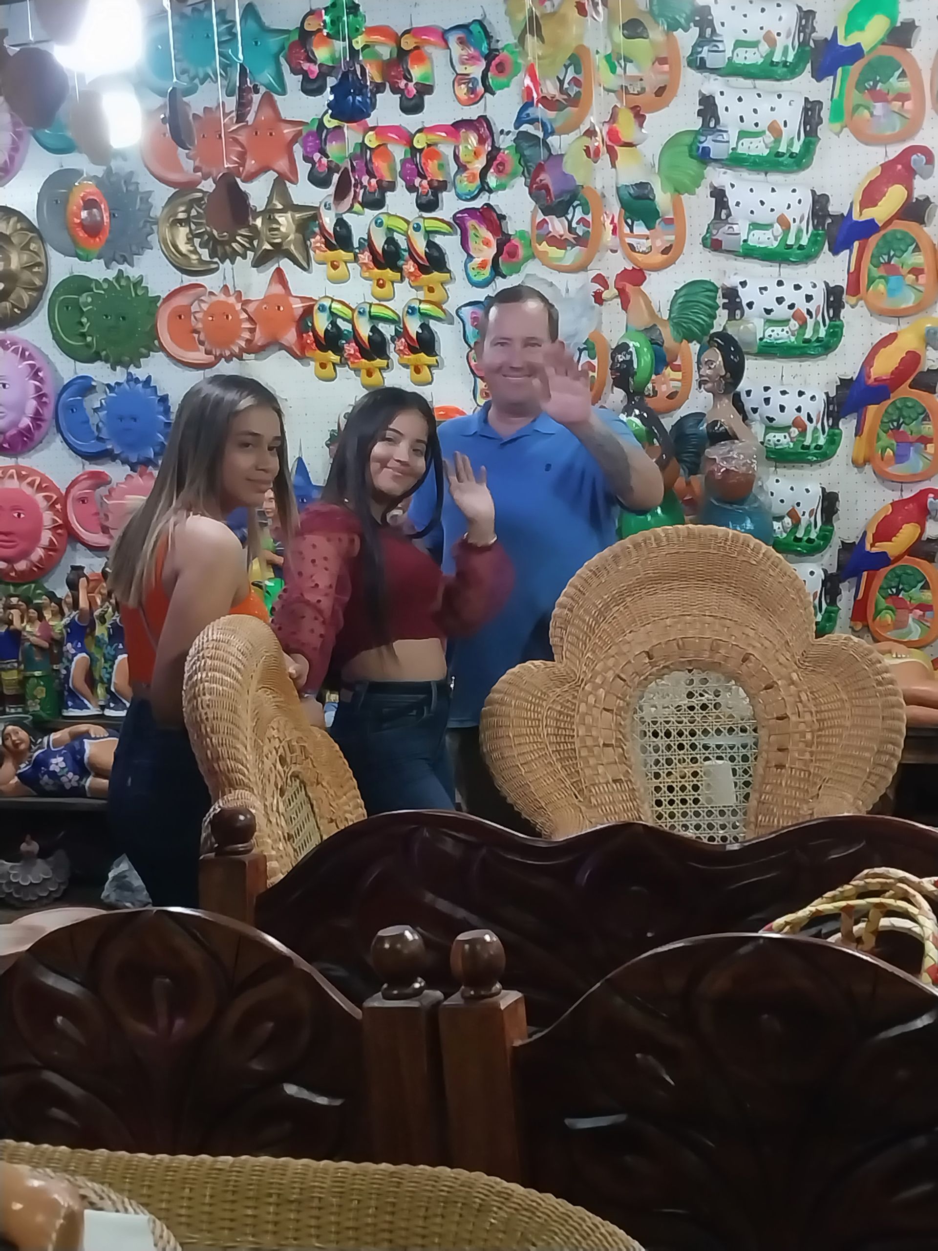 Three people pose in front of colorful wall art in a shop. One man waves, two women smile.