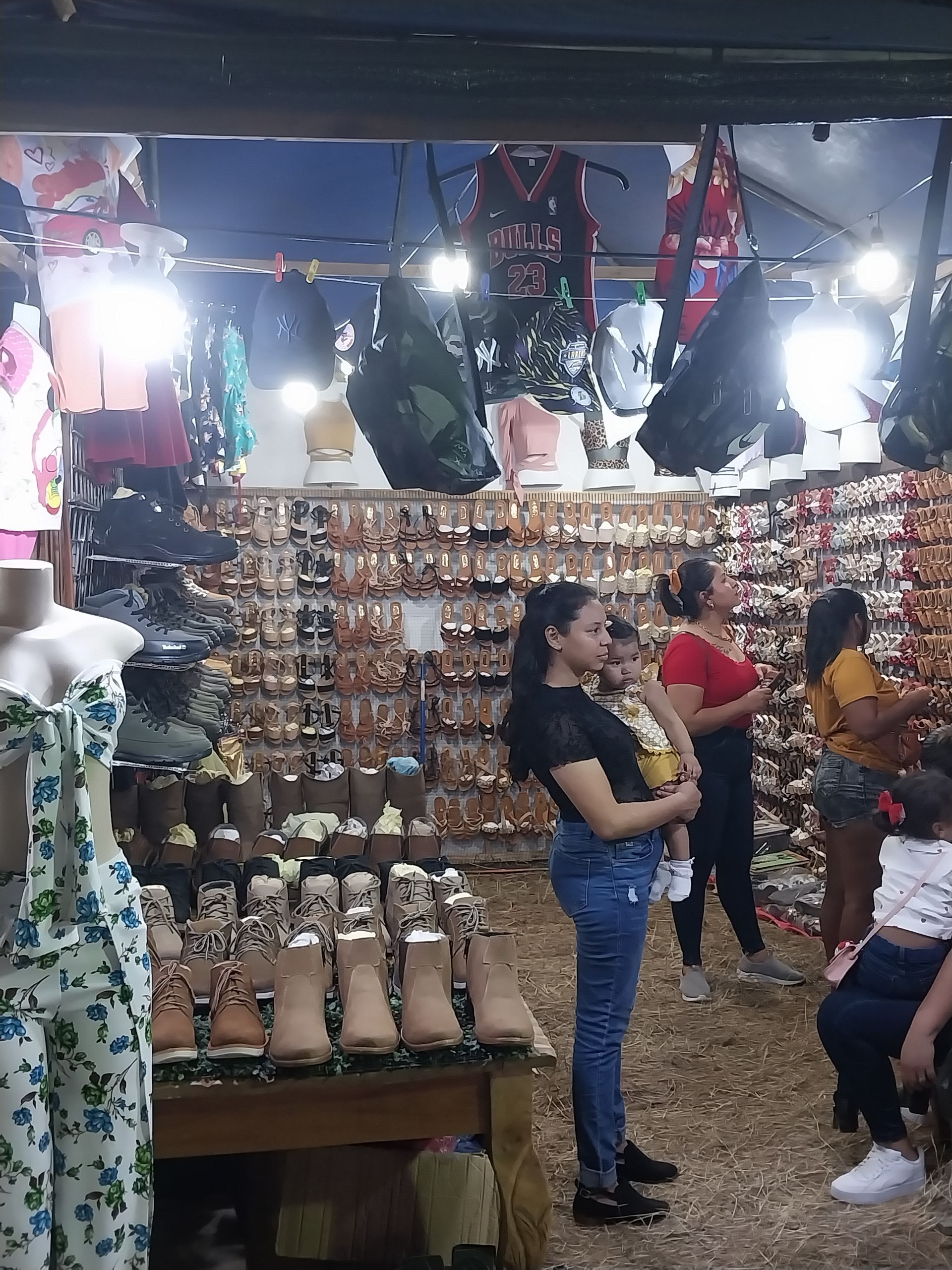 A woman holds a child in a crowded shoe and clothing store; shoppers browse merchandise.