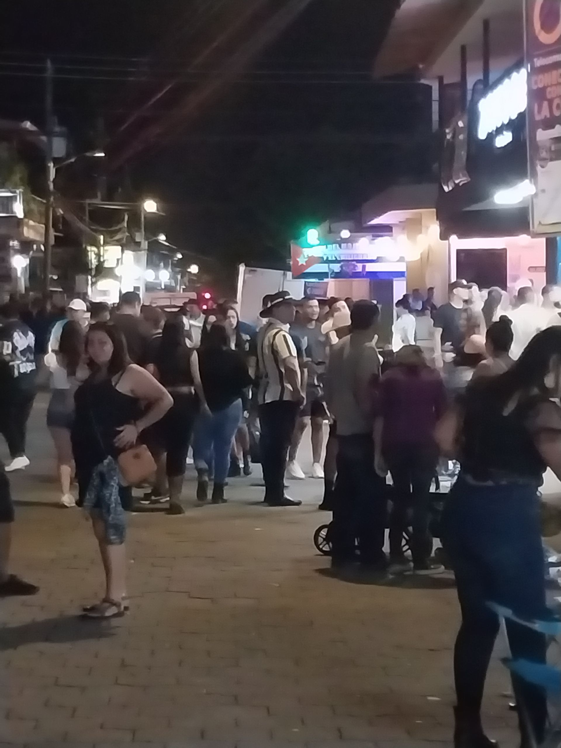 Nighttime street scene with crowd near lit storefronts. People are standing and socializing.