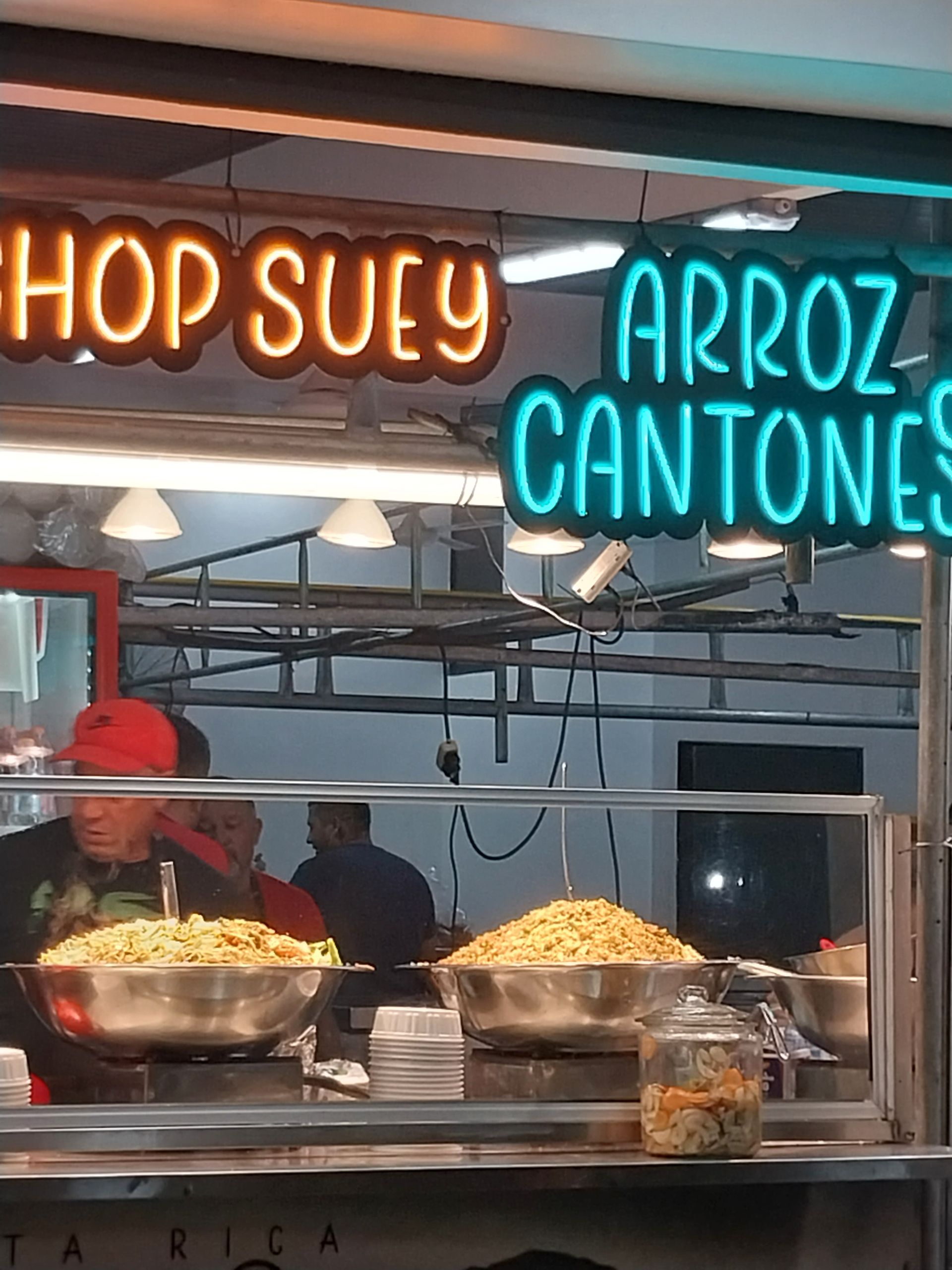 Neon signs for Chop Suey and Arroz Cantones over a food stall with two cooks serving food from large bowls.