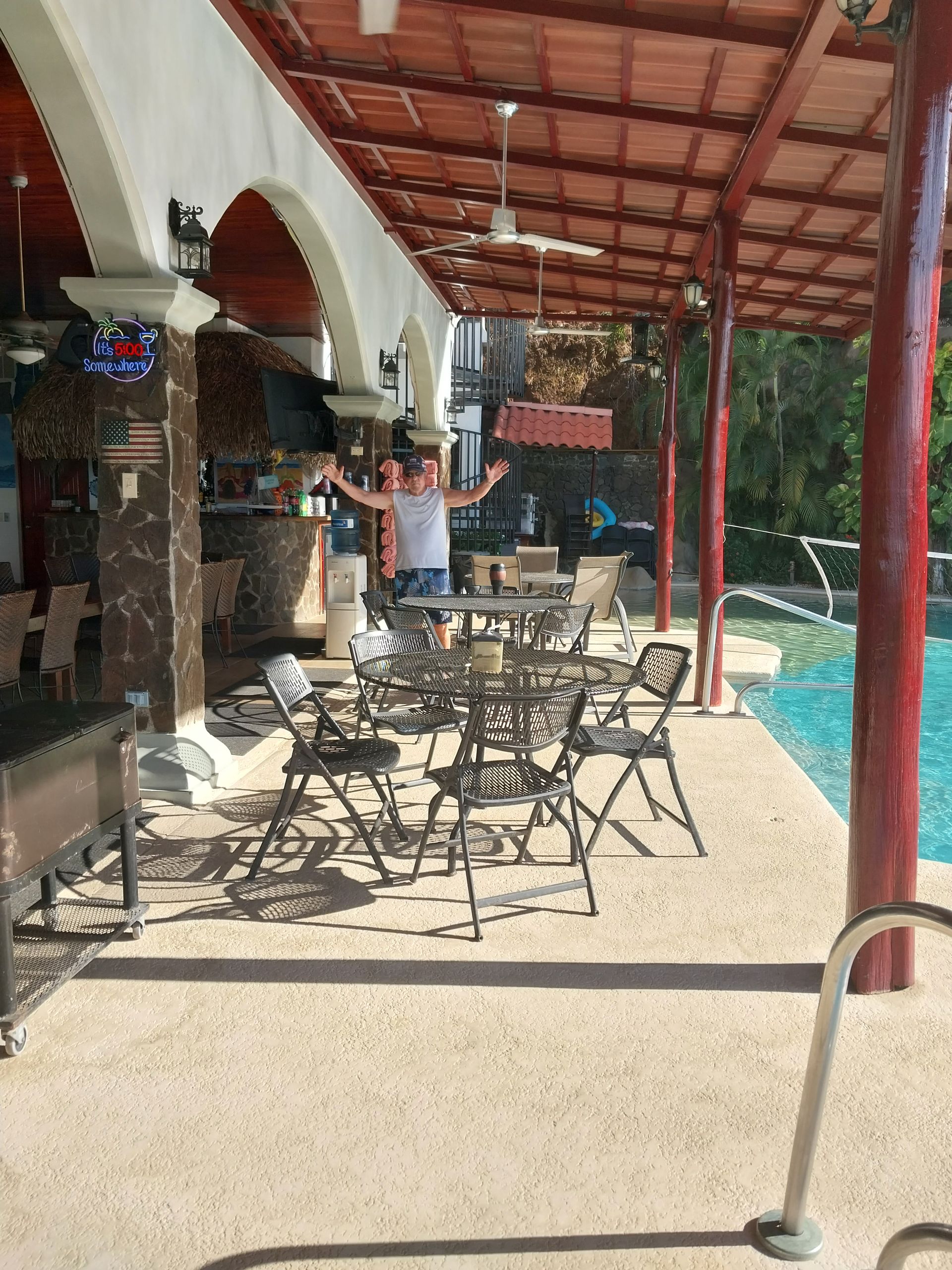 Man standing with arms open at outdoor bar by a pool. Tables and chairs are in front of the bar.