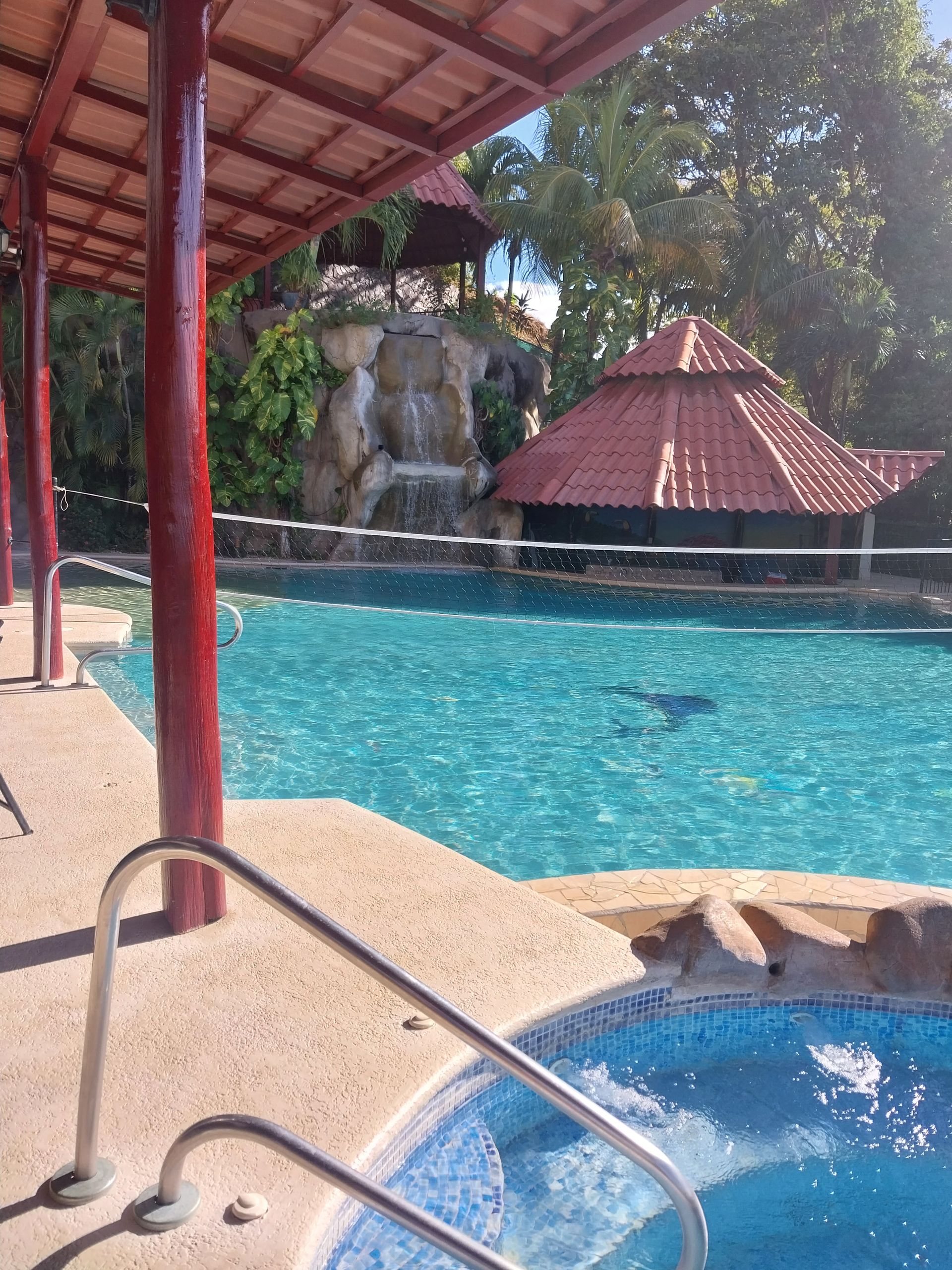 Pool with a waterfall, gazebo, and a red-roofed covered area. Sunny day, blue water.